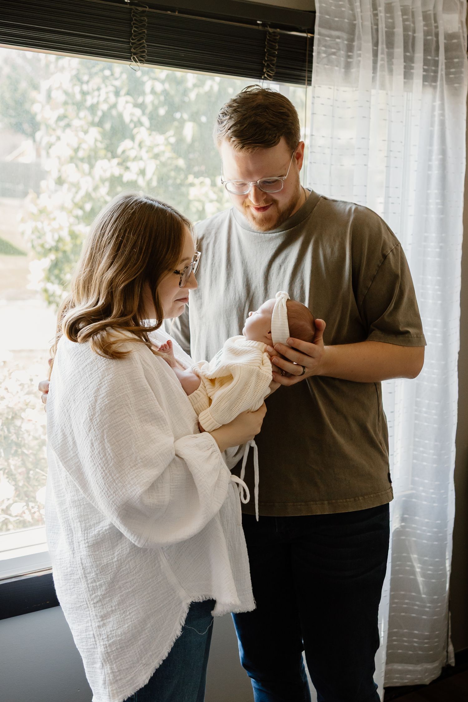New parents lovingly hold their swaddled newborn baby together by a bright window in a warm, intimate home setting.