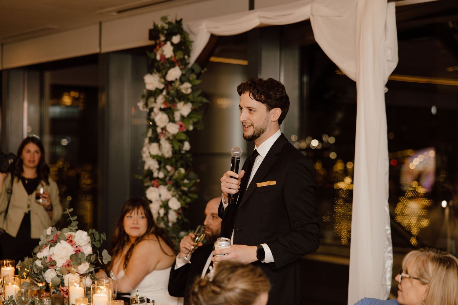 A person gives a wedding speech under an elegant floral arch with candlelit tables and evening ambiance in view.