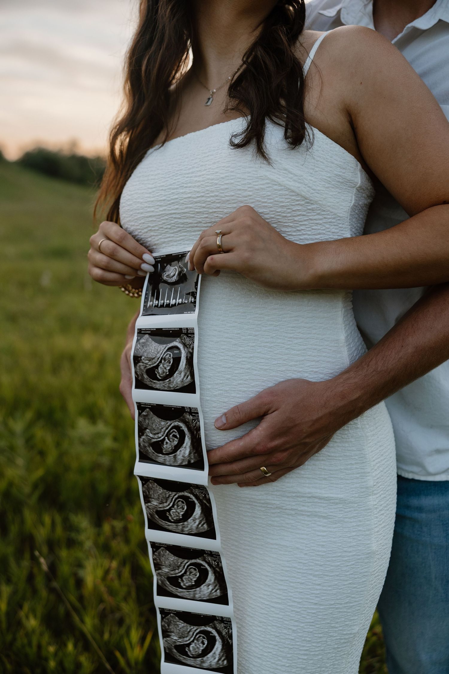 Couple holding ultrasound photos during sunset maternity photoshoot in field.