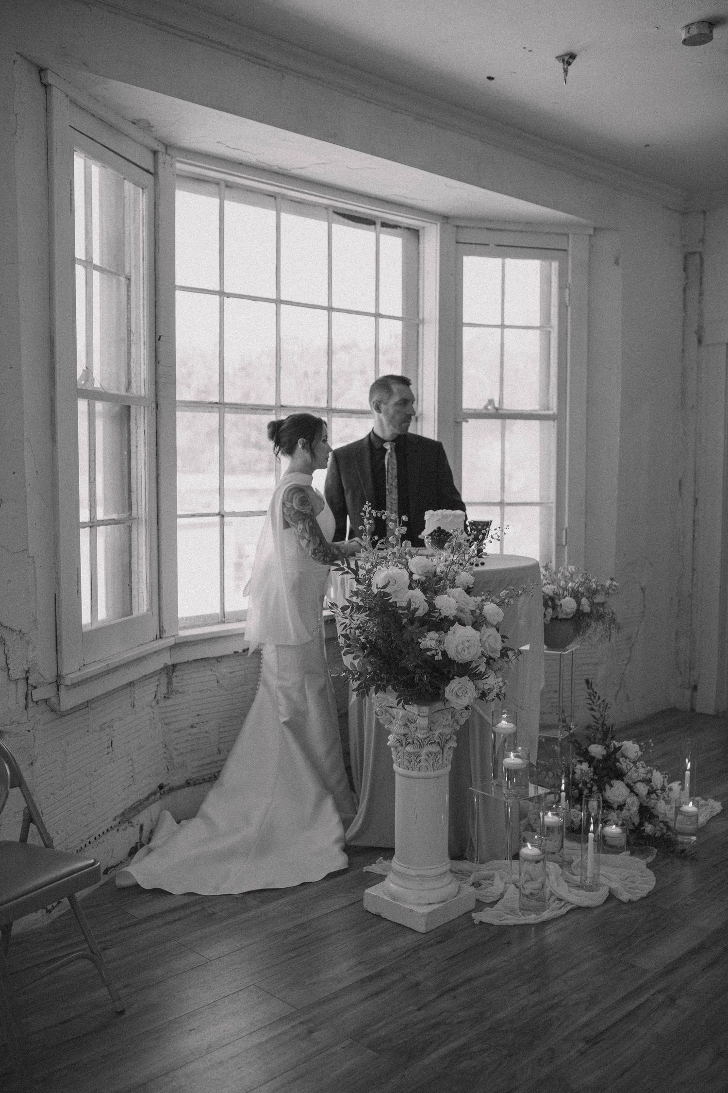 A black and white wedding scene with a couple standing by a window beside elegant floral arrangements and candlesticks.