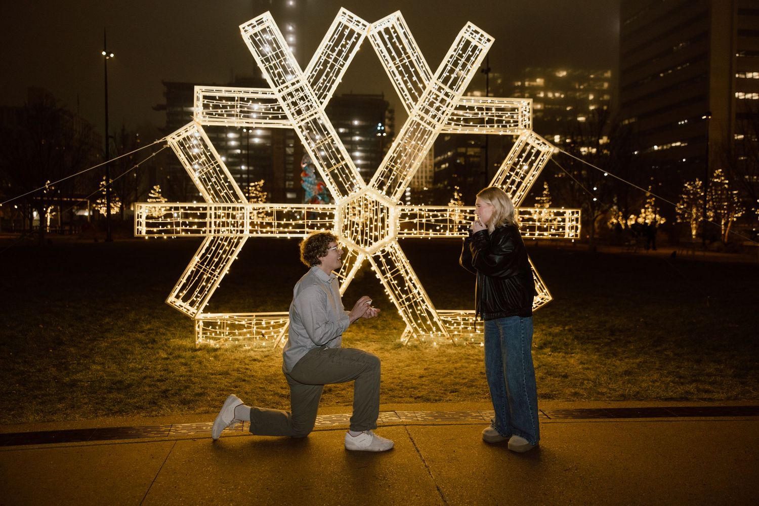 A couple stands in front of a large illuminated snowflake decoration at night.