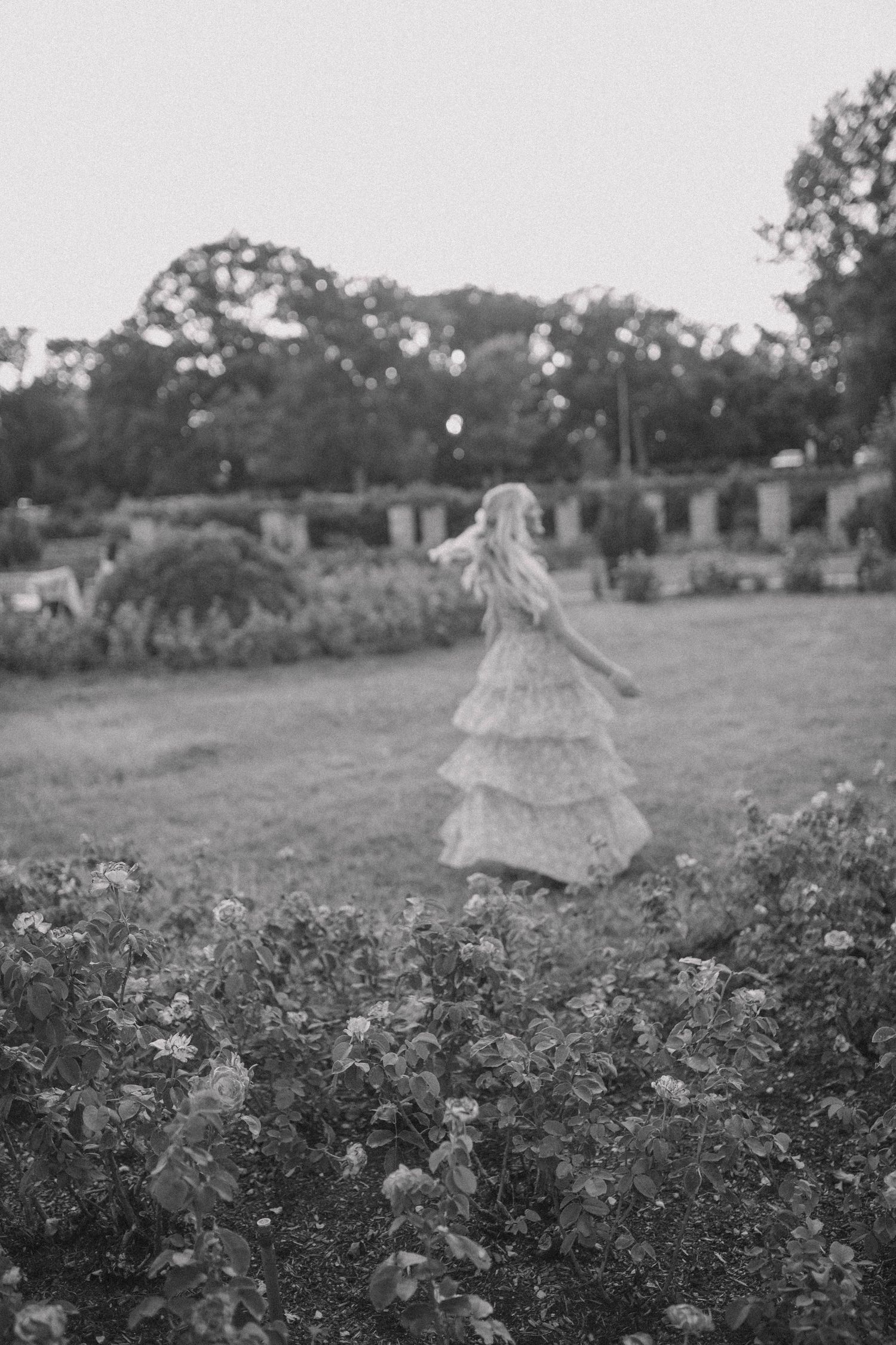 A black and white photo of someone twirling in a tiered dress among garden flowers.
