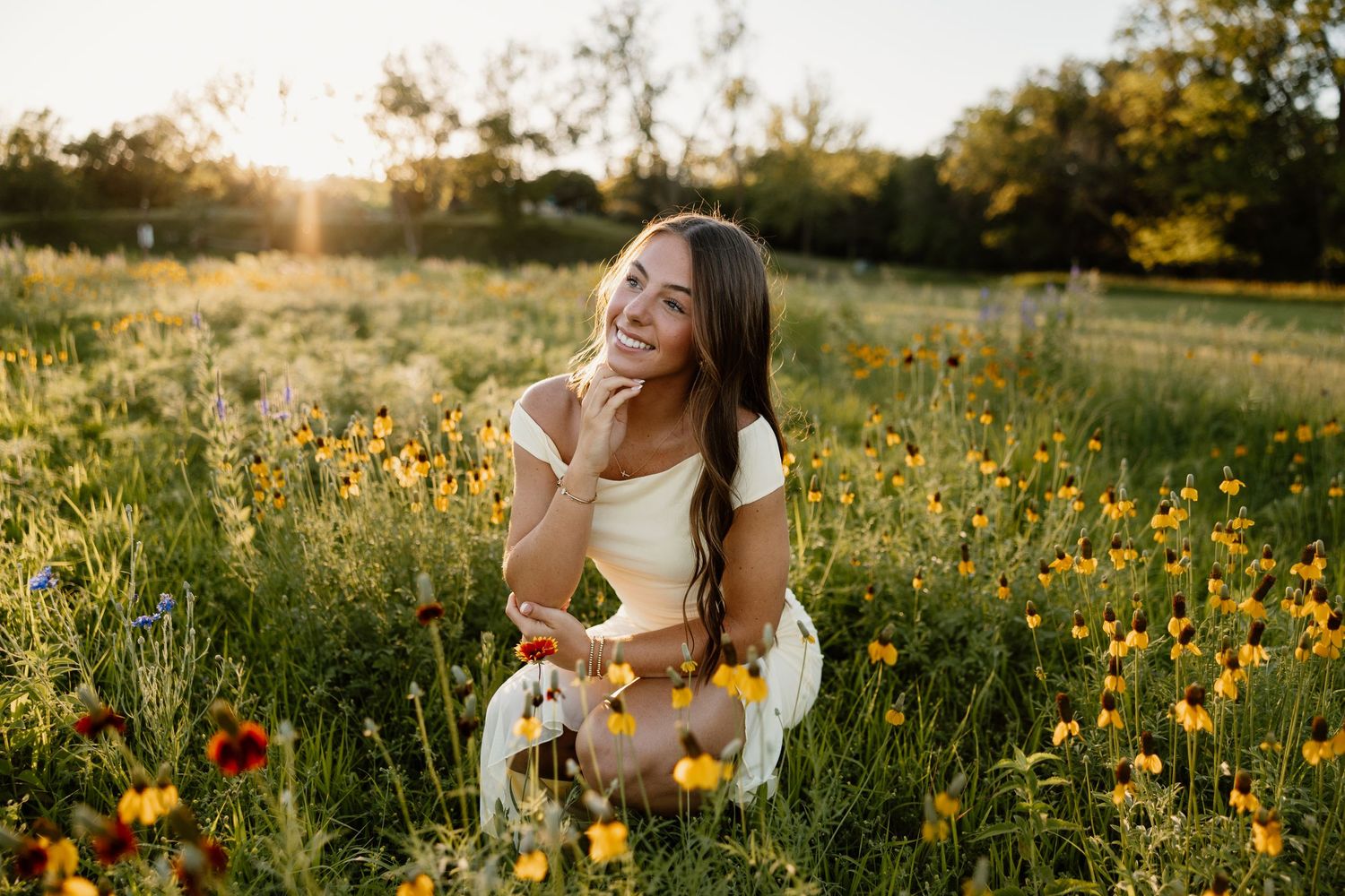 Young woman smiling in a sunlit wildflower meadow, wearing a white dress, sitting among colorful blooms at golden hour.