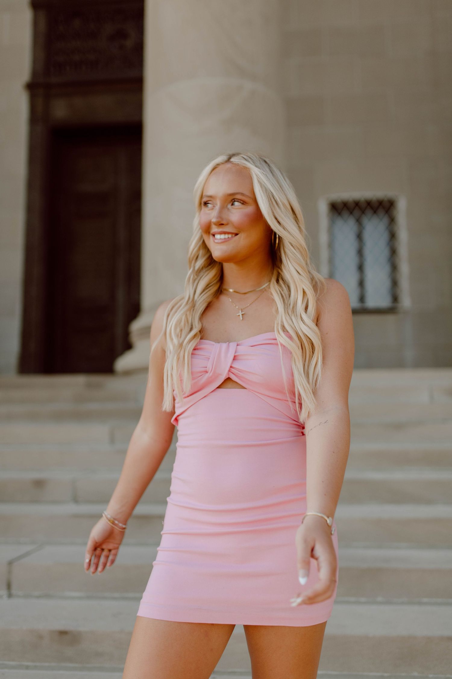 Young woman in a pink mini dress poses confidently on outdoor stone steps.