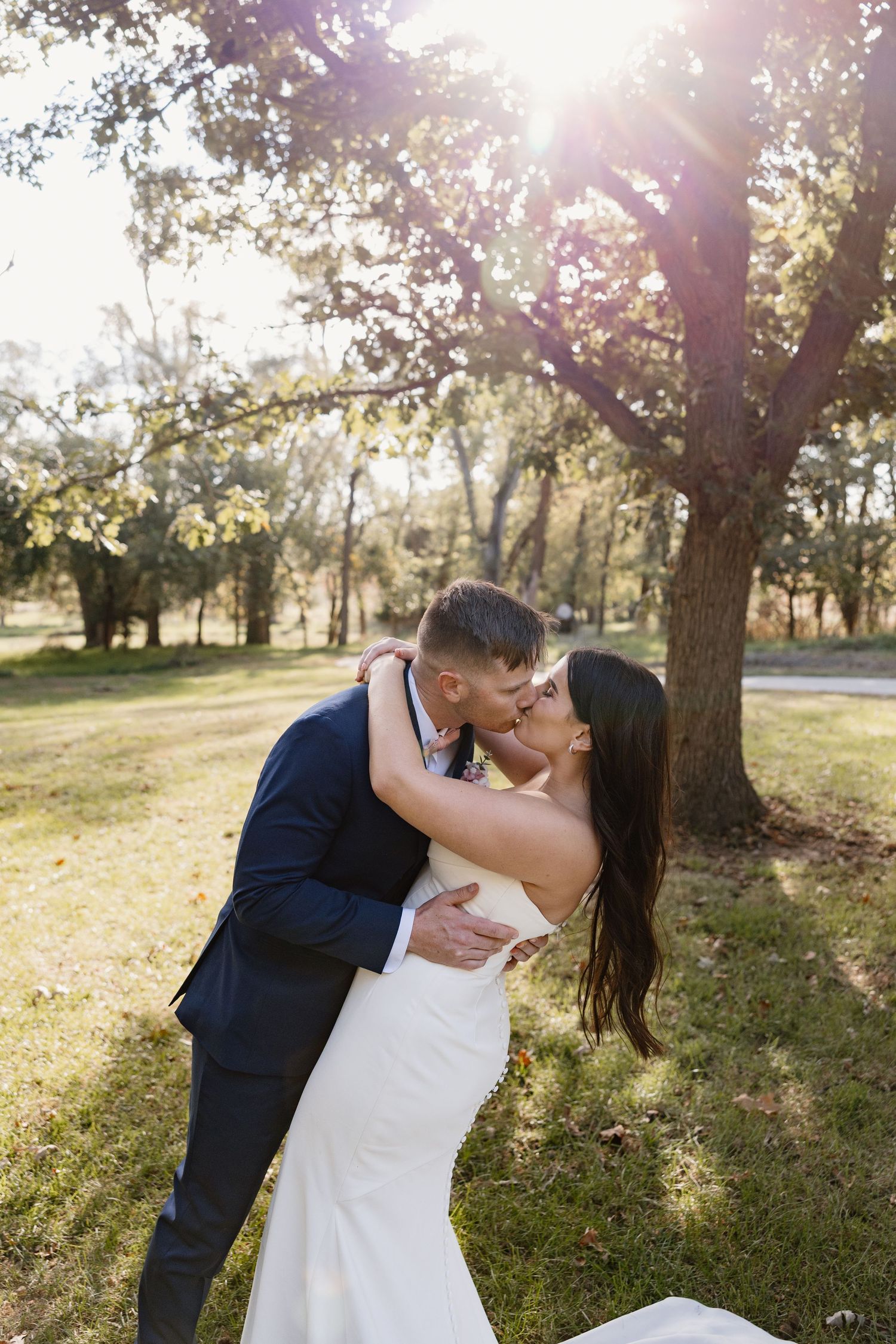 Romantic wedding couple embraces and kisses in a sunlit park with trees in the background.