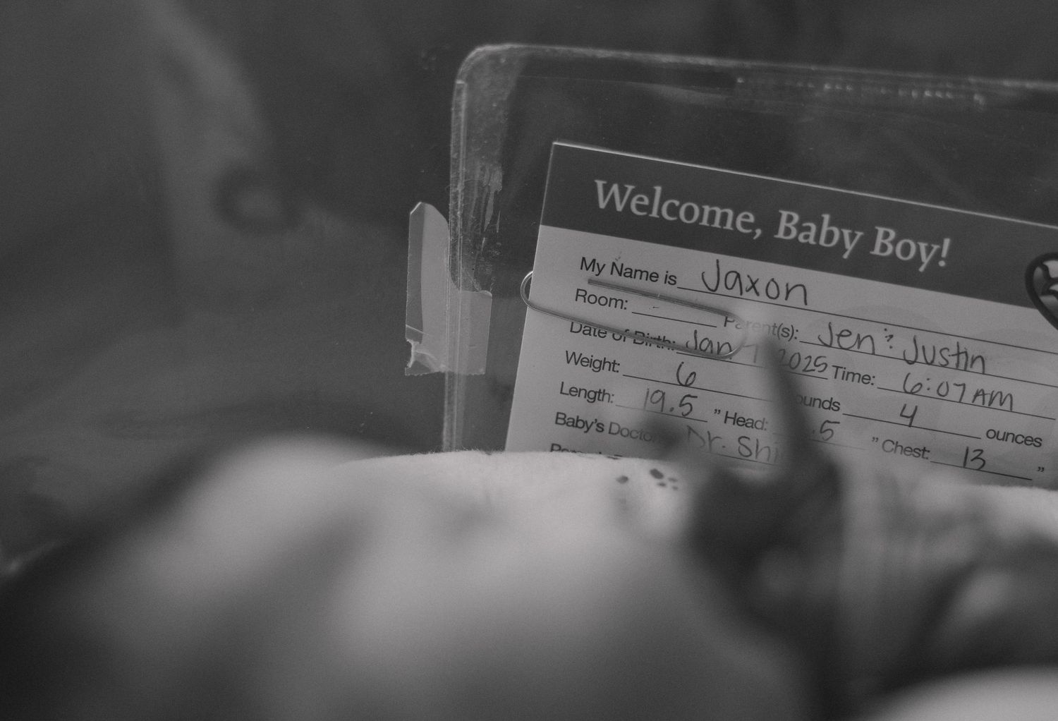 A newborn baby rests in hospital beneath a 'Welcome Baby Boy' card showing birth details in black and white.