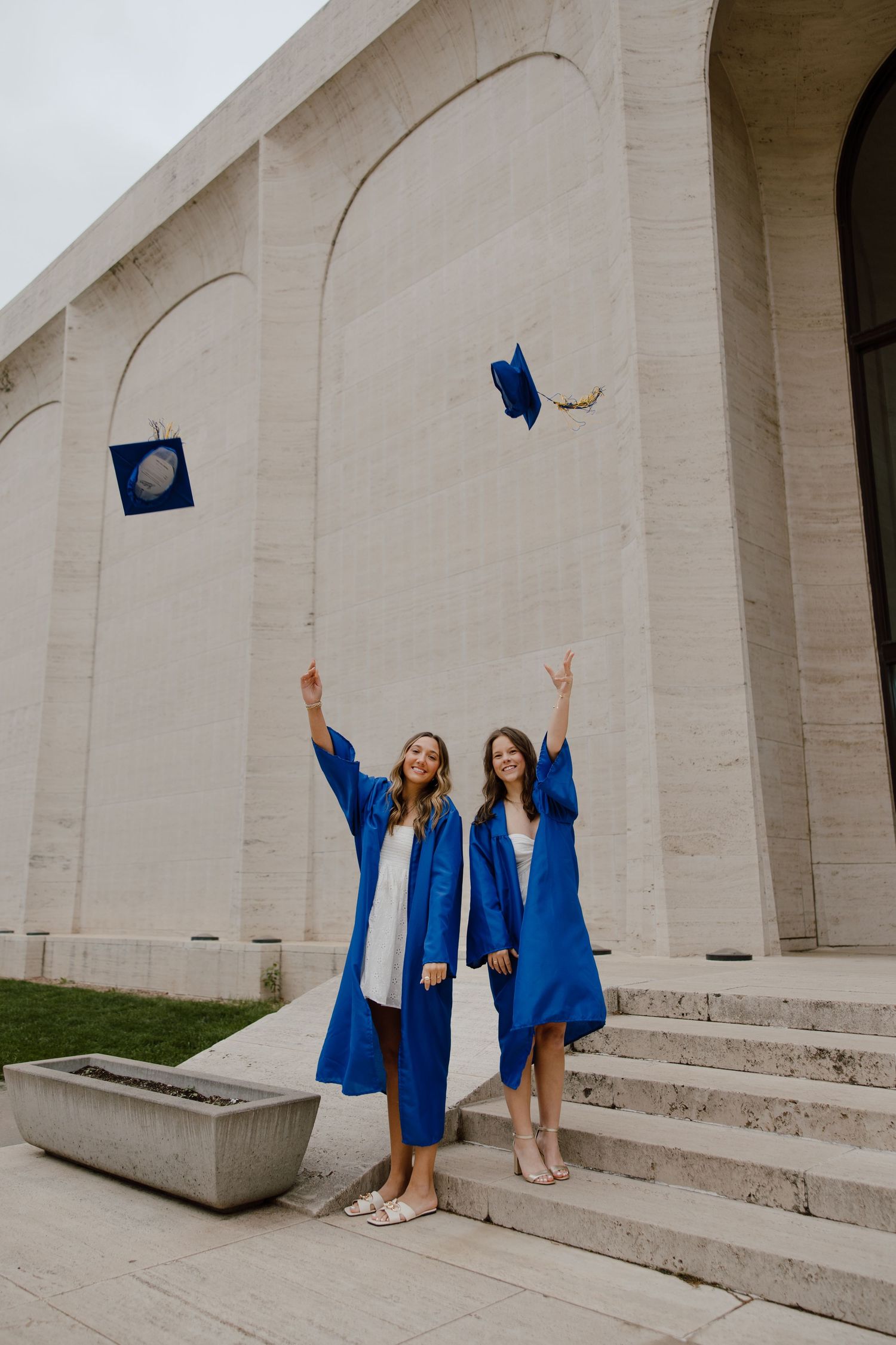 Two graduates in blue caps and gowns pose for photos in front of a white stone building.