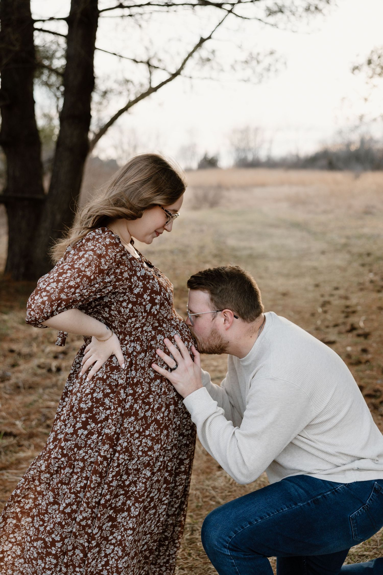 A man kneels to kiss a pregnant woman's belly outdoors in a field during a maternity photo session in Omaha Nebraska.