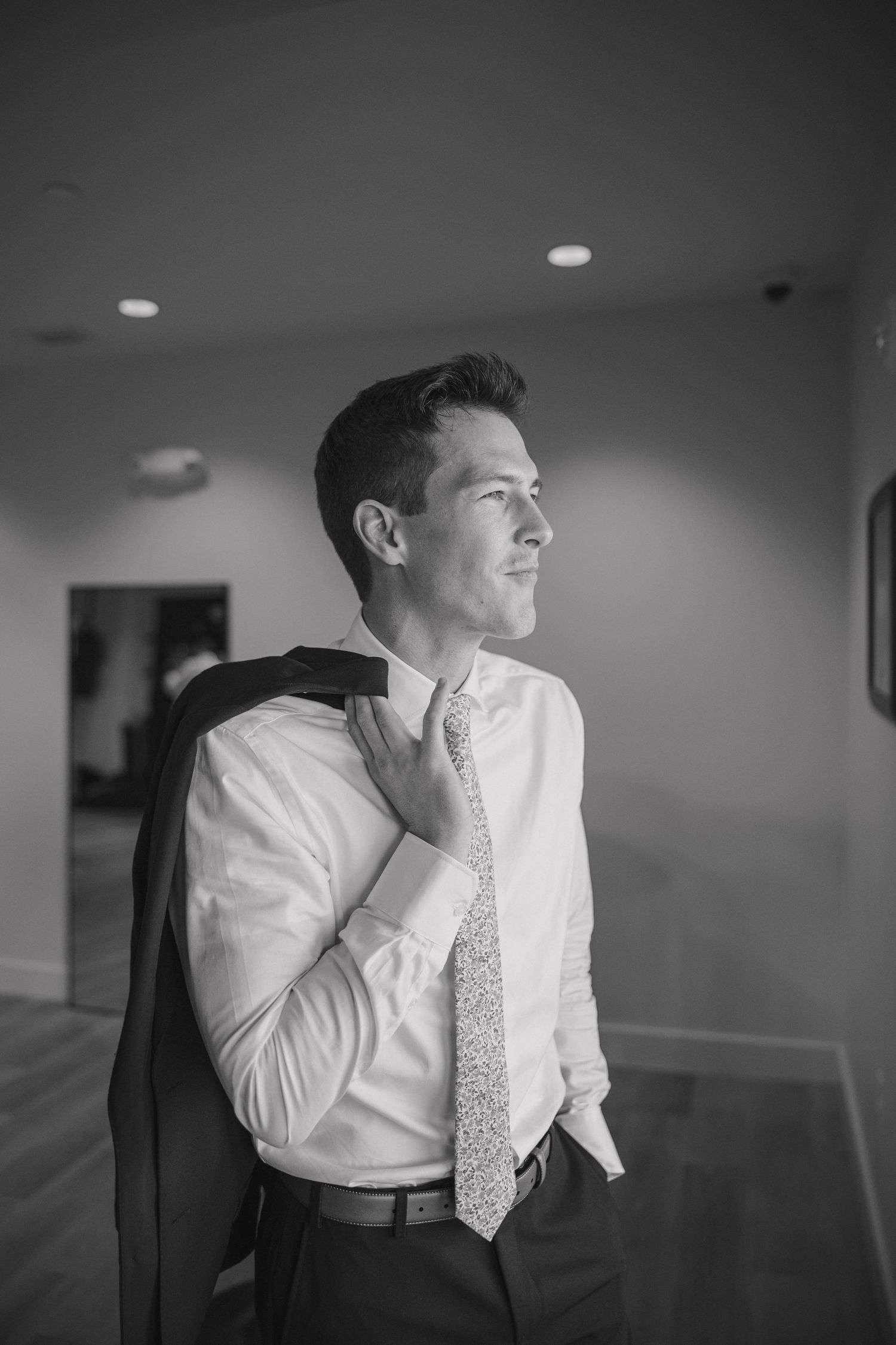 Black and white photo of a well-dressed man in a white shirt and tie, holding a suit jacket over his shoulder indoors.