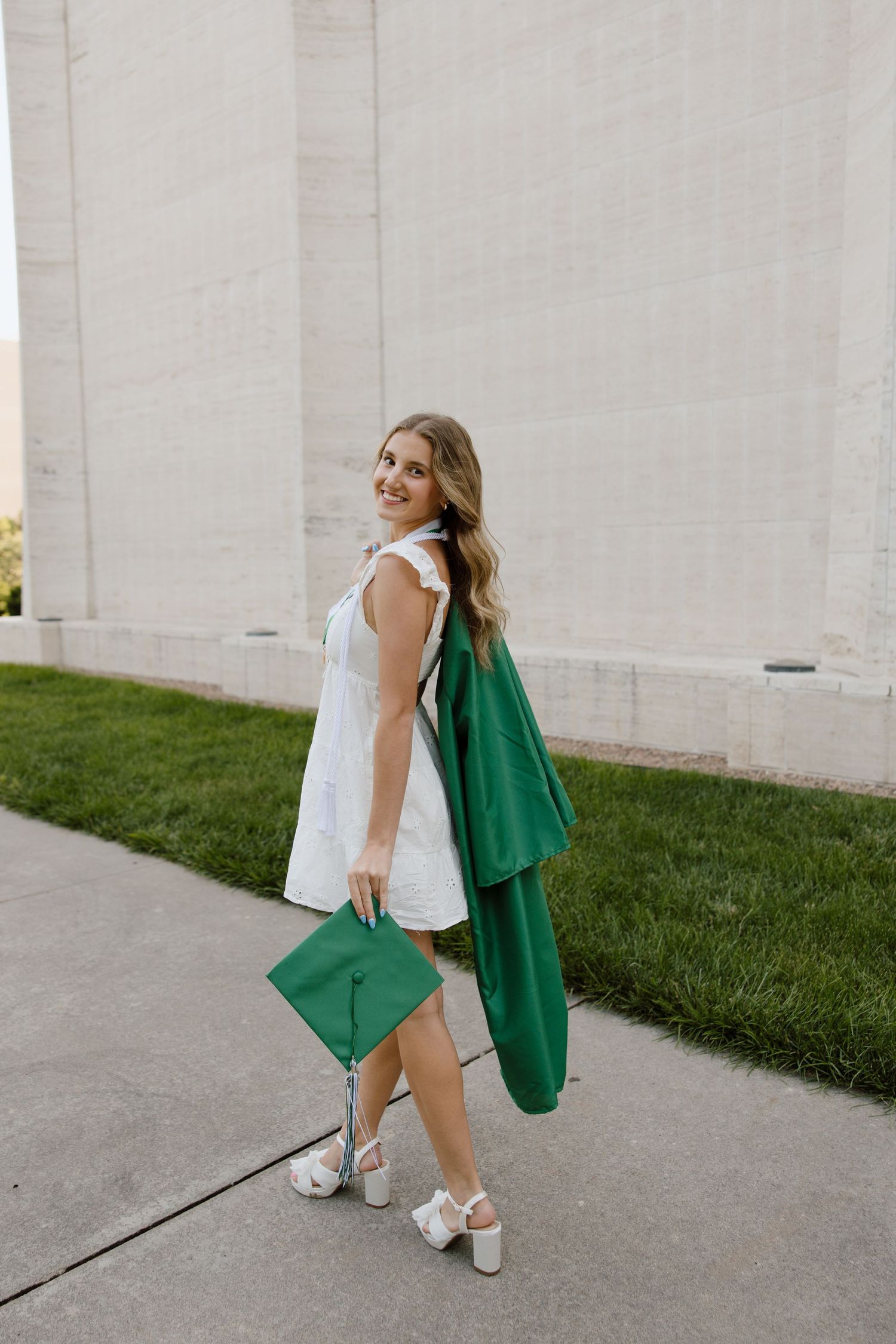 Graduate in white dress holding green graduation gown poses near concrete building.