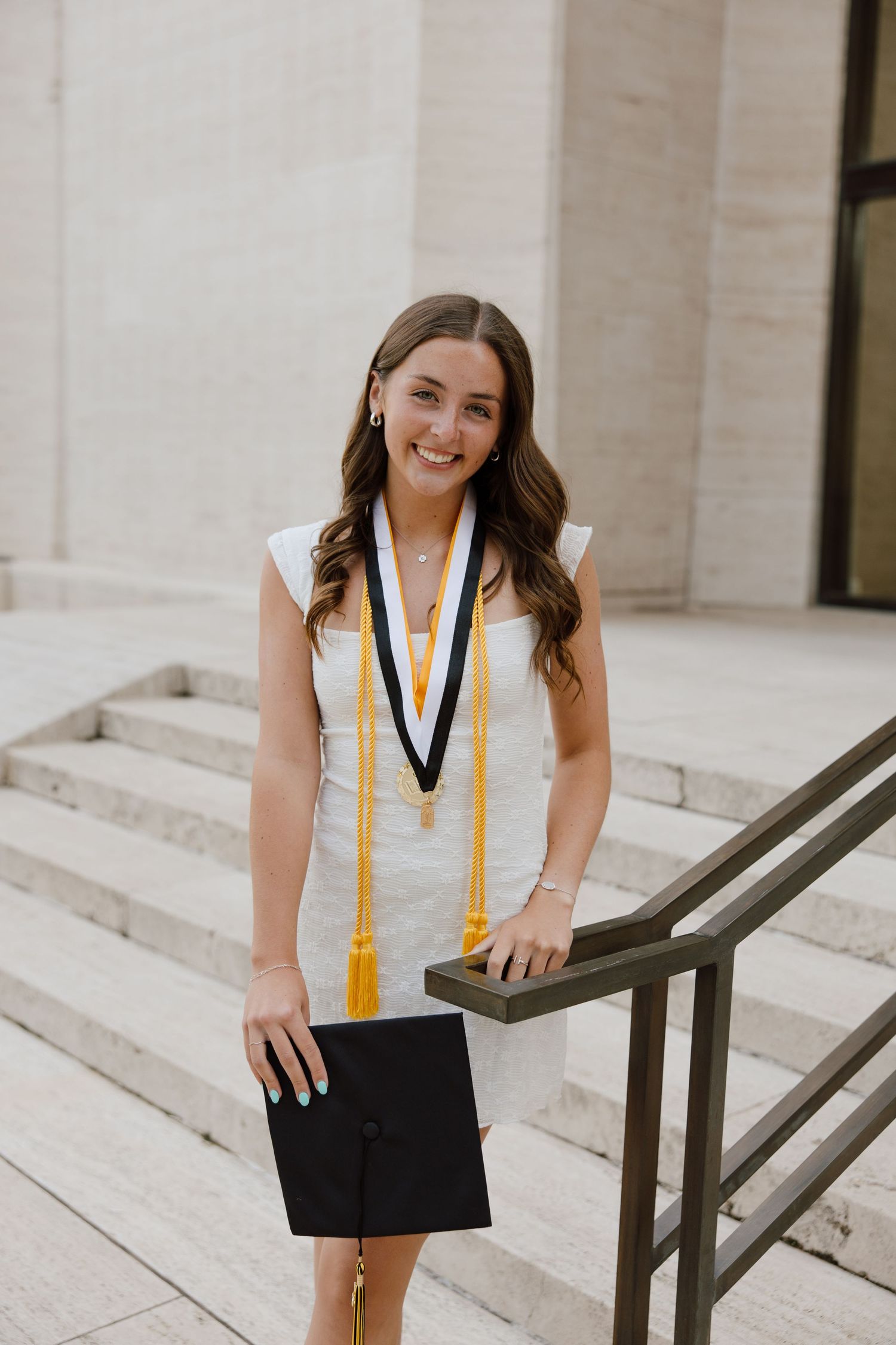 Graduate in white dress holding graduation cap on stone steps with honor cords and medal.
