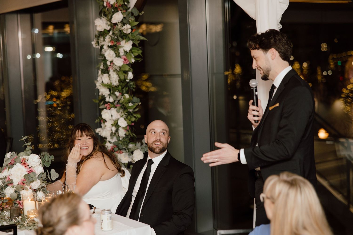 Wedding guests share a joyful candid moment during reception toasts with elegant floral decor in the background.