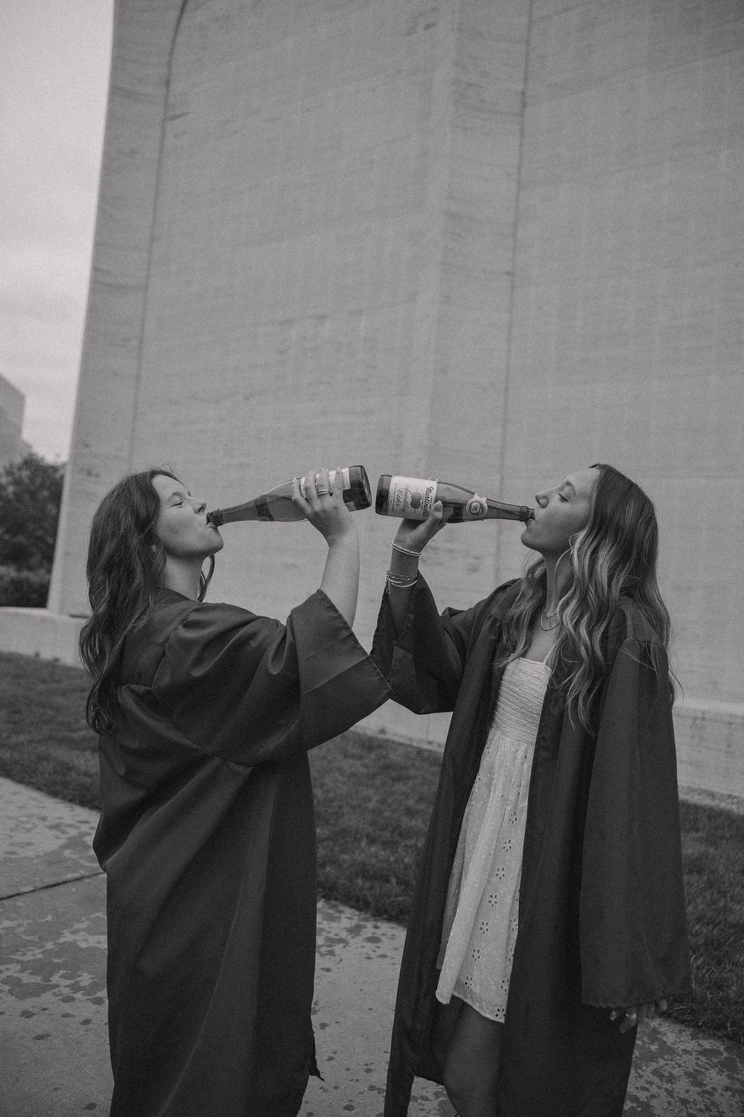 Two graduates in black robes sharing a celebratory drink outdoors.