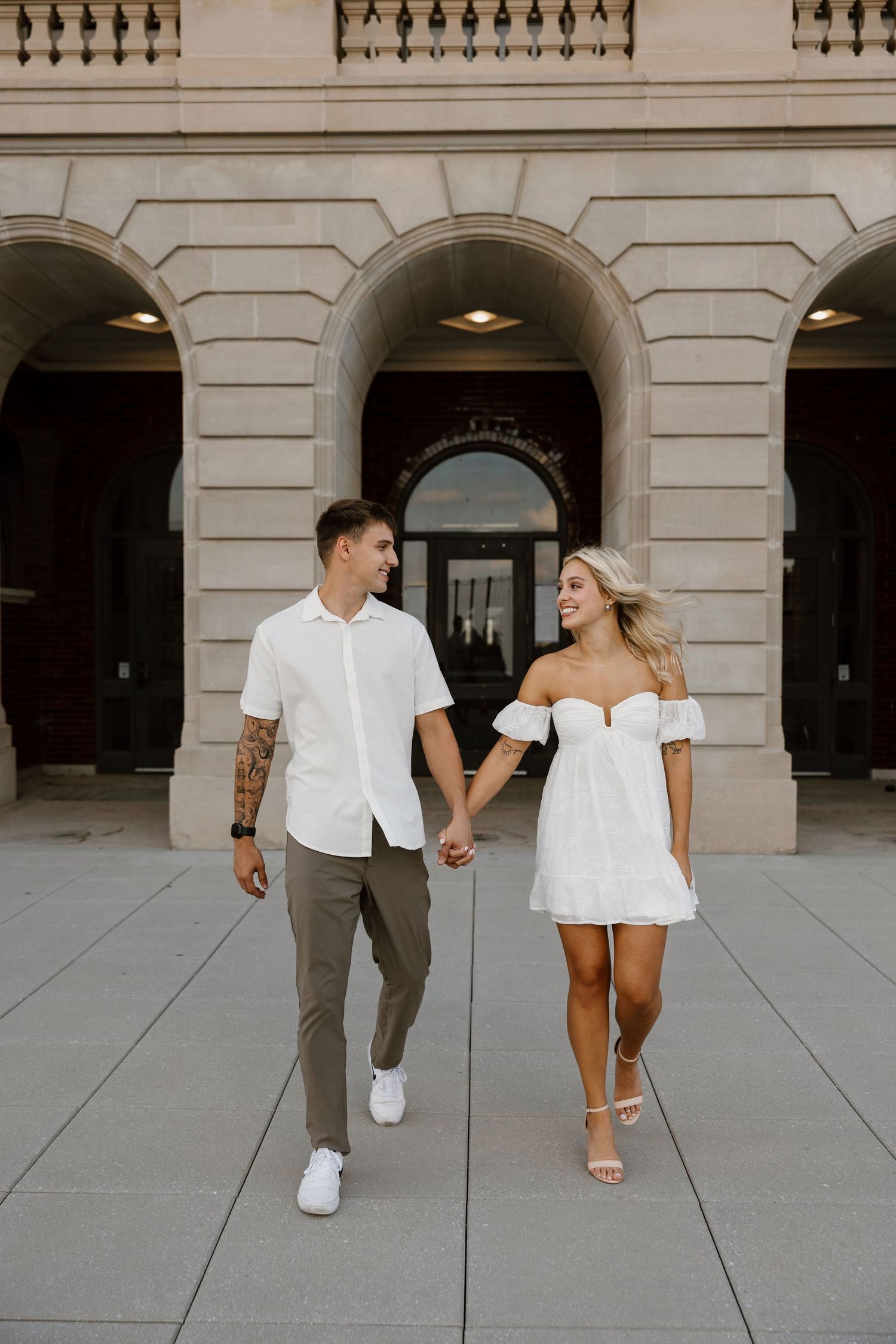 A couple walks hand in hand along a grand stone building with arched entrances.