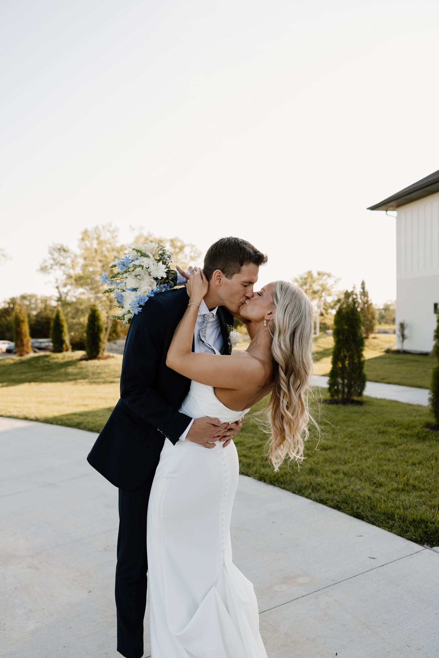 A bride and groom share a romantic kiss outdoors on their wedding day, surrounded by lush greenery near a white barn.