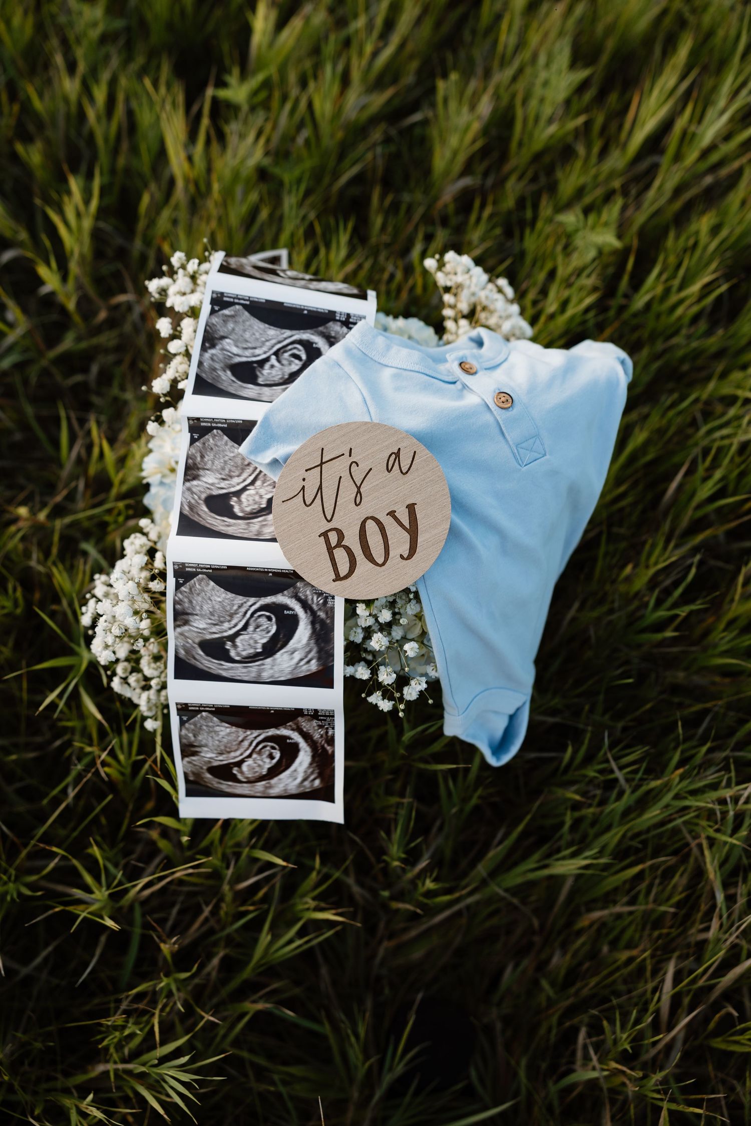 Ultrasound photos and a light blue baby onesie arranged with white flowers on grass announce it's a boy.