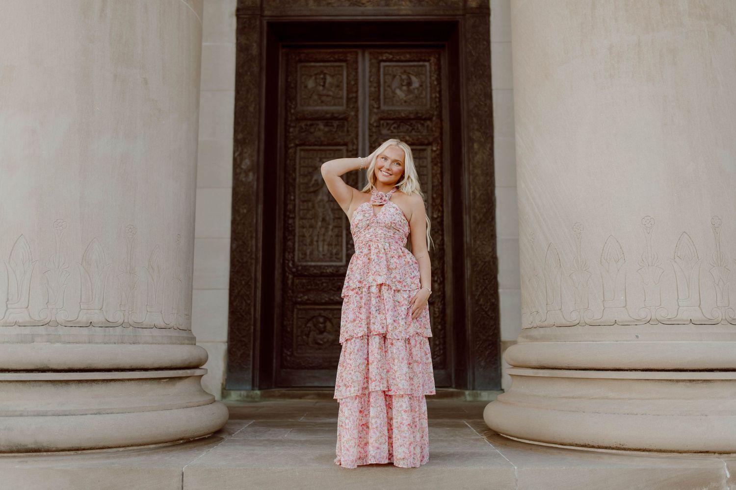 Woman in long floral dress poses between classical columns near ornate wooden door.