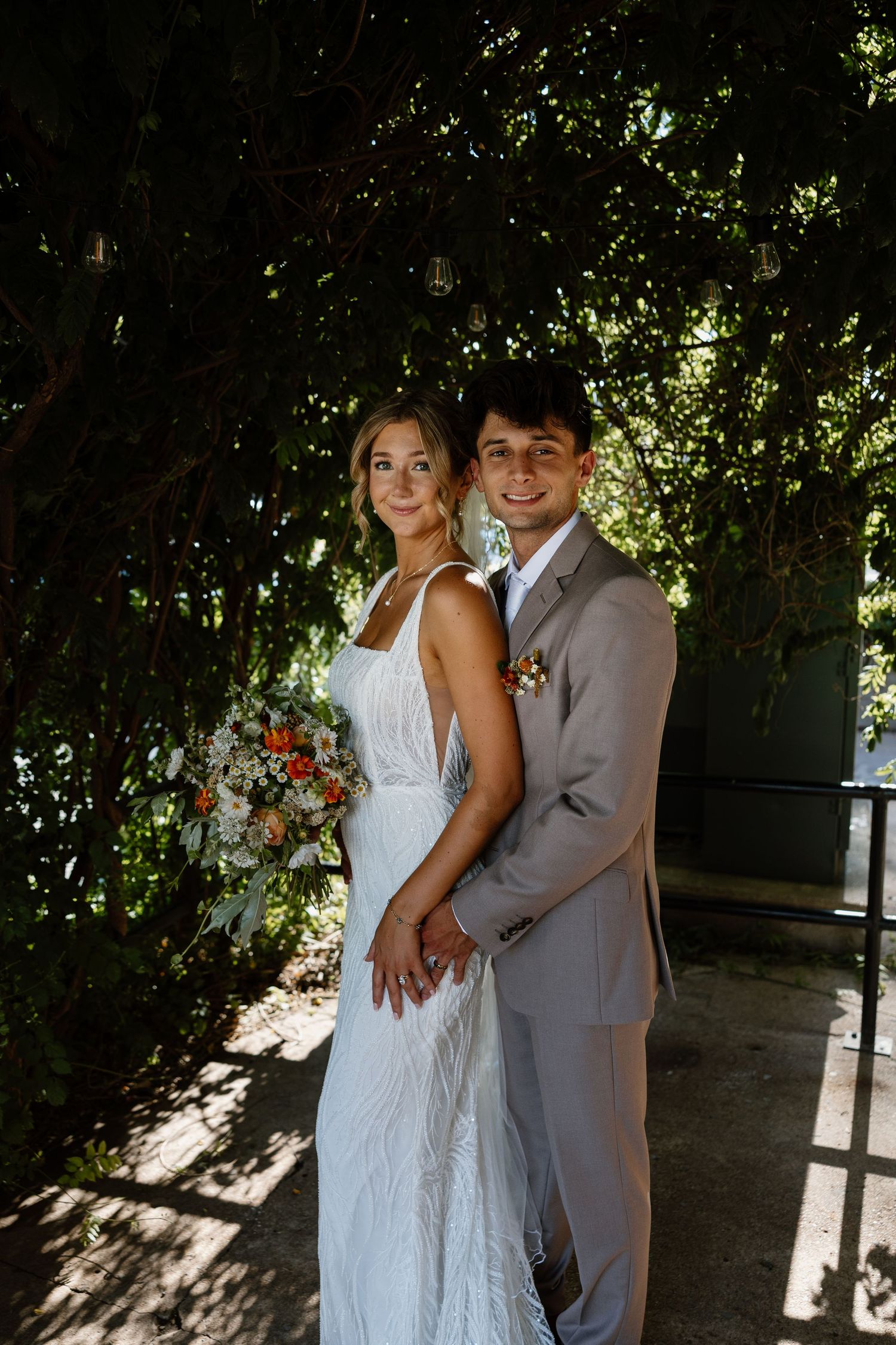 Wedding couple poses in dappled sunlight against lush garden backdrop.