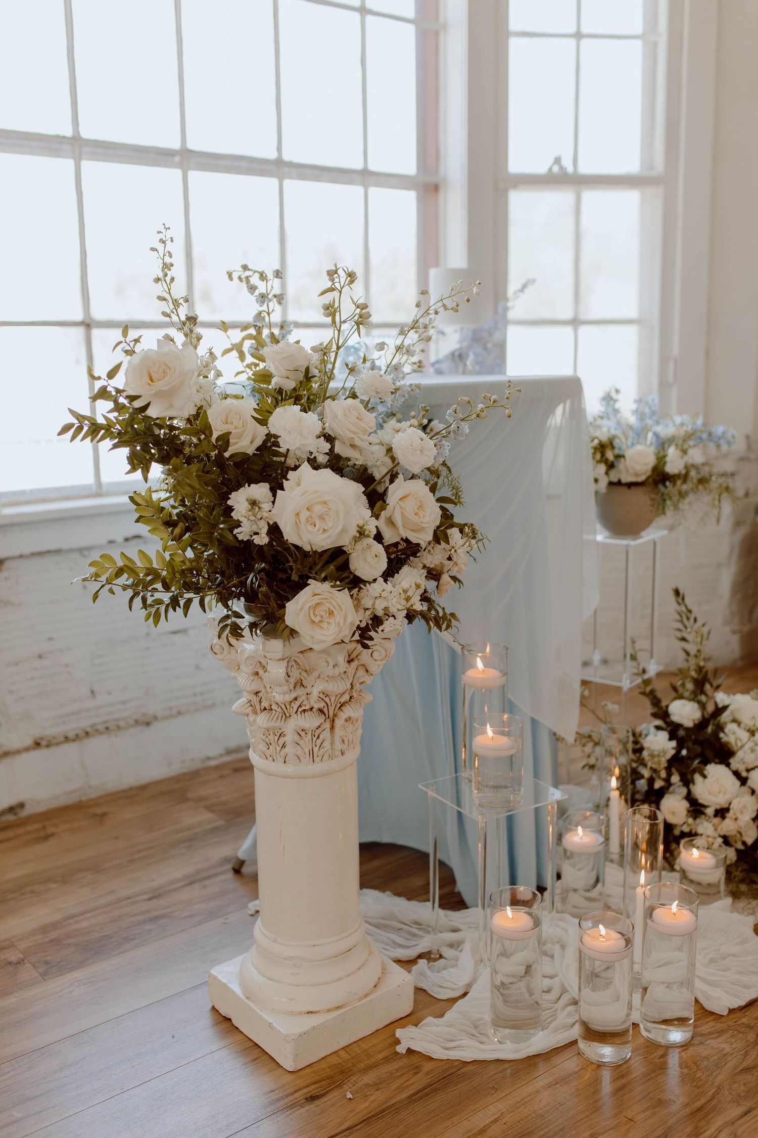 White roses and greenery arranged in an elegant vase on a pillar, with glowing candles creating a romantic wedding atmosphere.