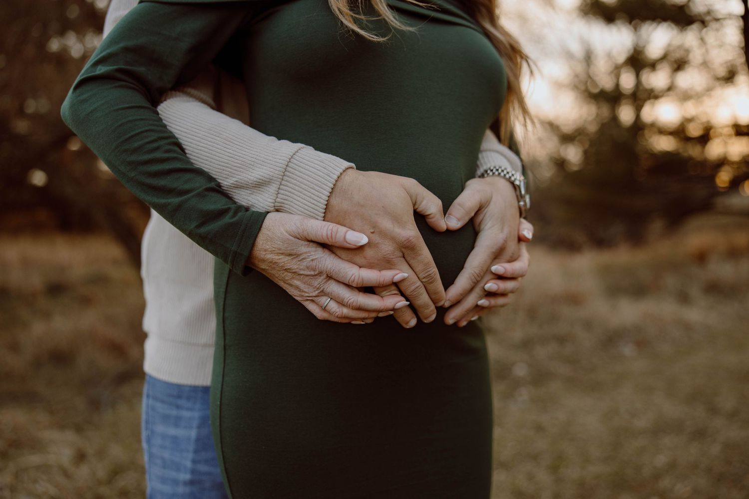 Hands forming a heart shape over a pregnant belly in a green dress at sunset.