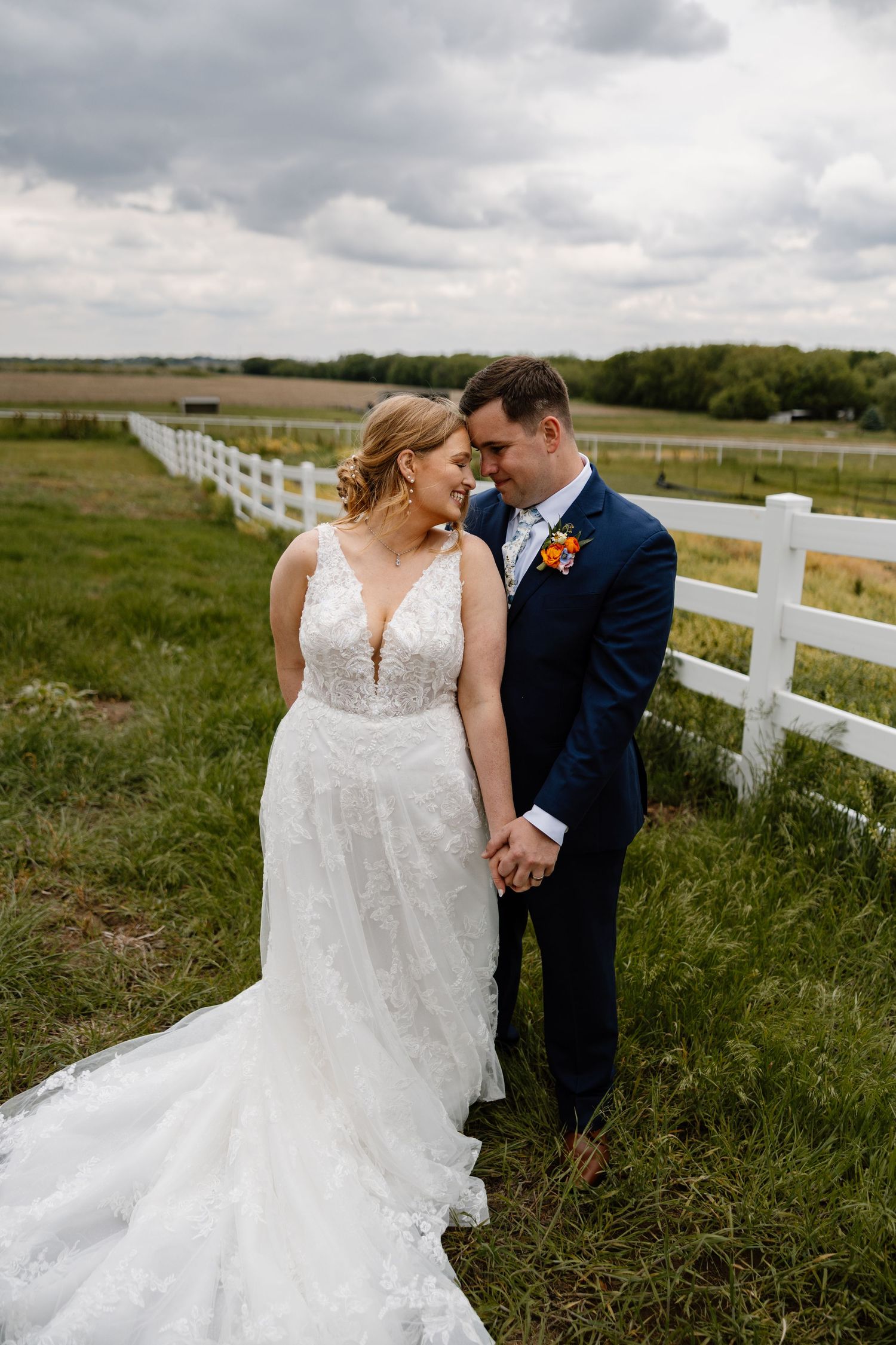 Newlyweds share tender moment by white fence with bride in lace gown and groom in navy suit.
