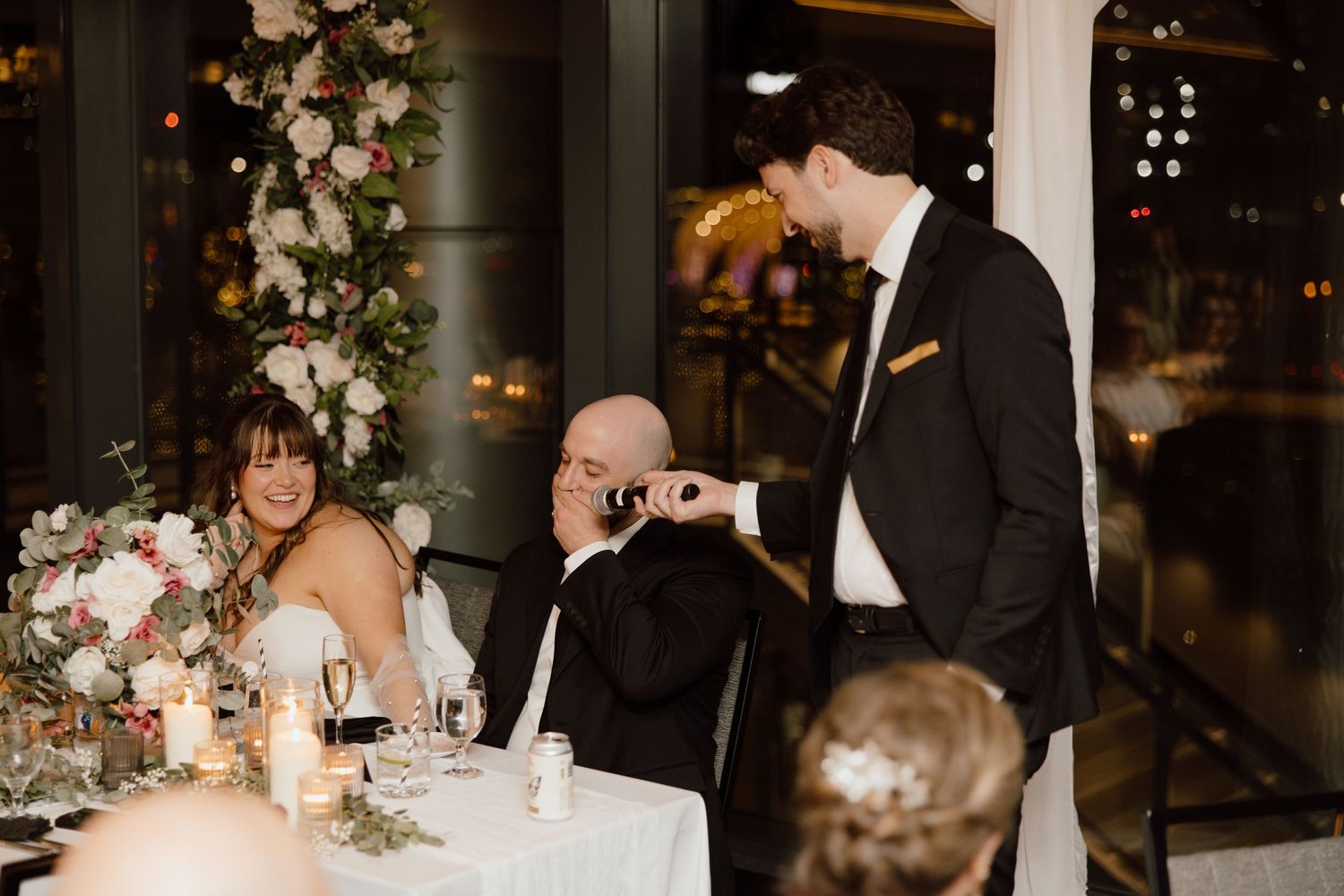Wedding reception guests react during candid moment at an elegant dinner table decorated with floral arrangements and candles.