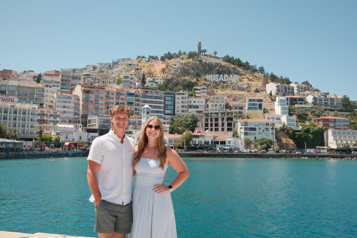 Couple posing by turquoise harbor with colorful hillside town and Kudasai sign in background on sunny day.
