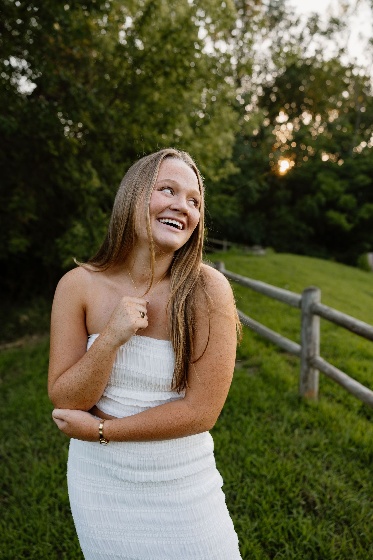 A person in a white dress poses outdoors against a rustic wooden fence at sunset with joyful expressions.