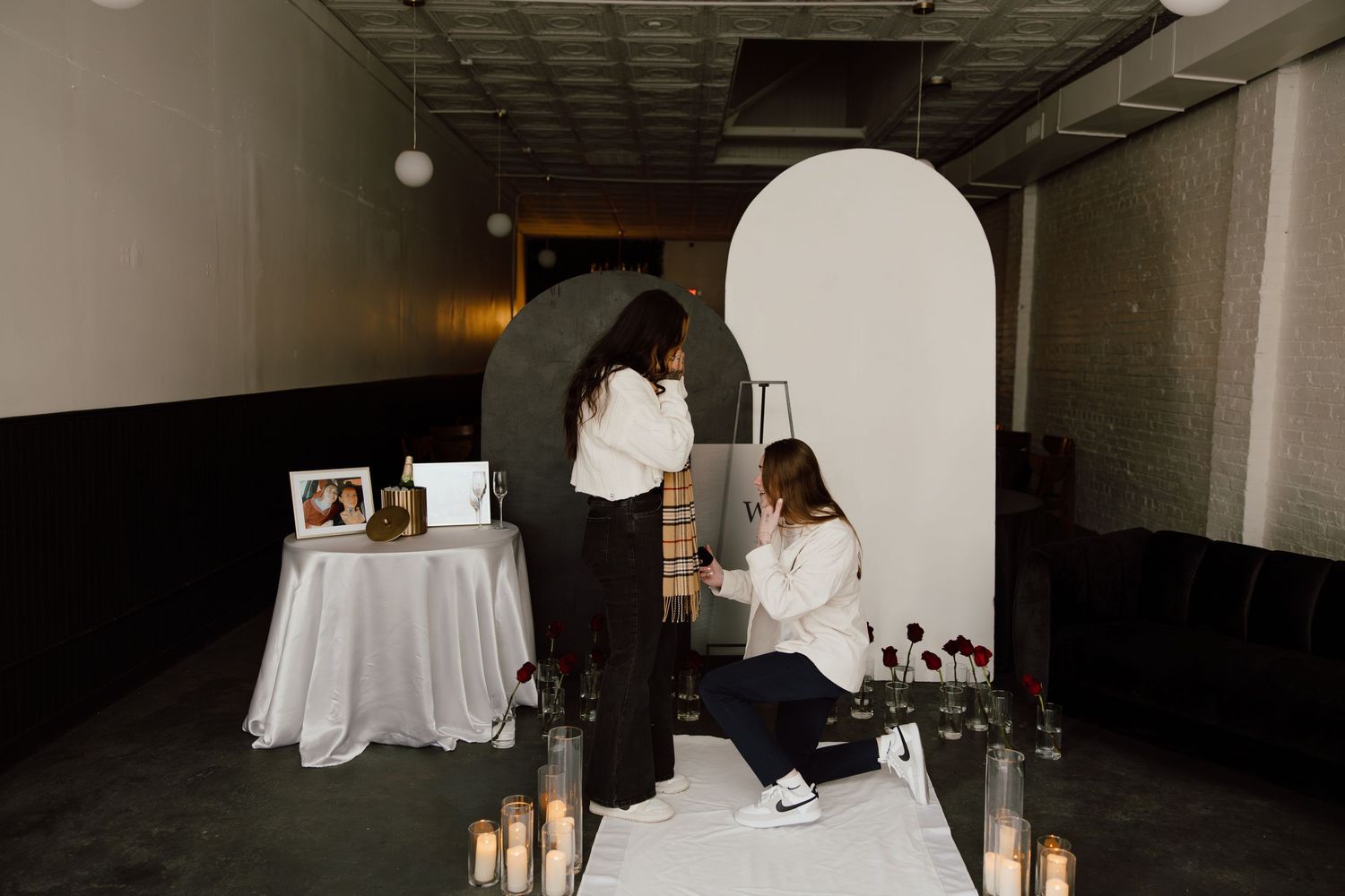 Two people embrace in a dimly lit indoor space with white walls.