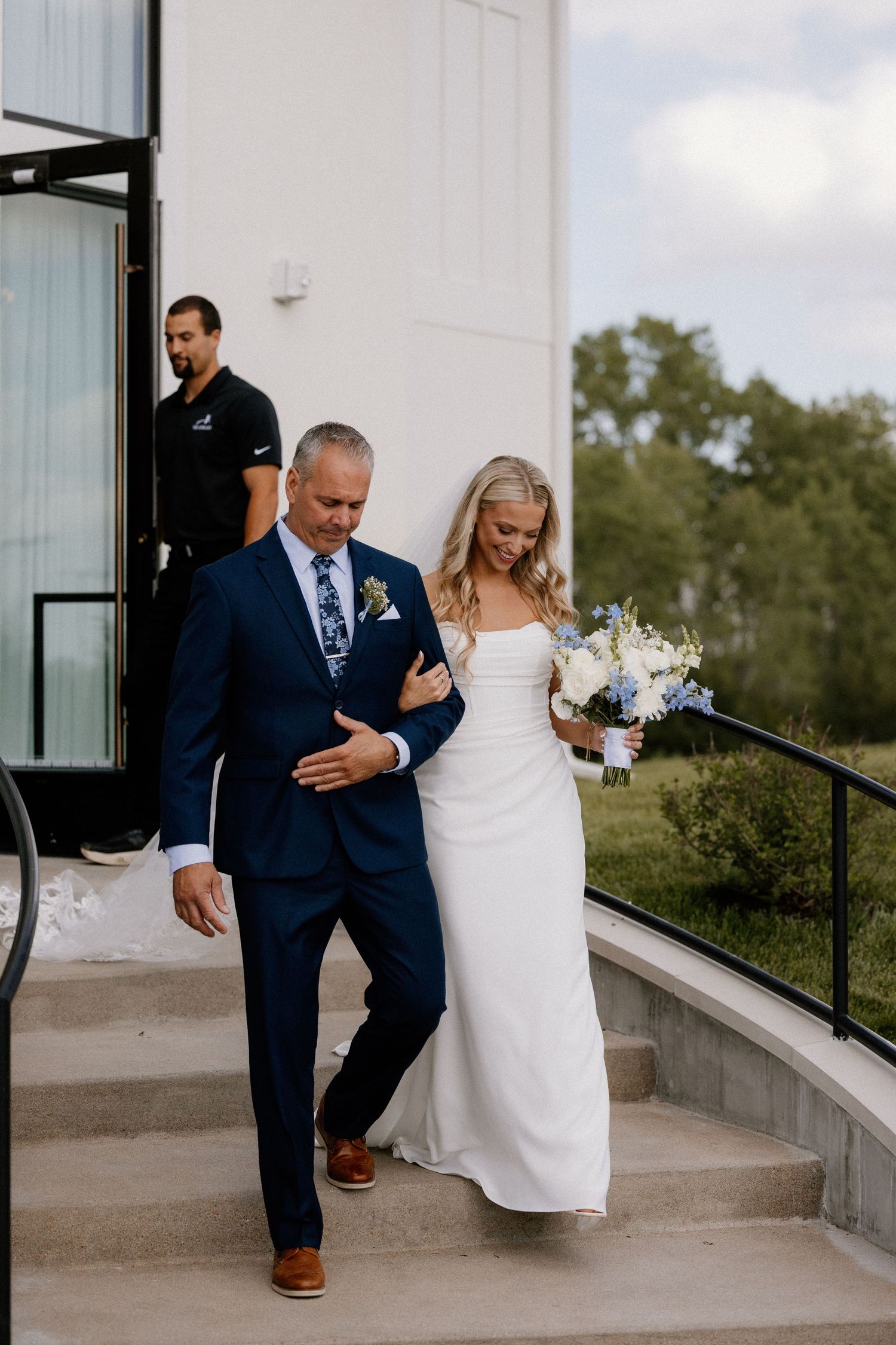 Bride in white gown holds bouquet while walking down steps with man in navy suit at outdoor wedding venue.