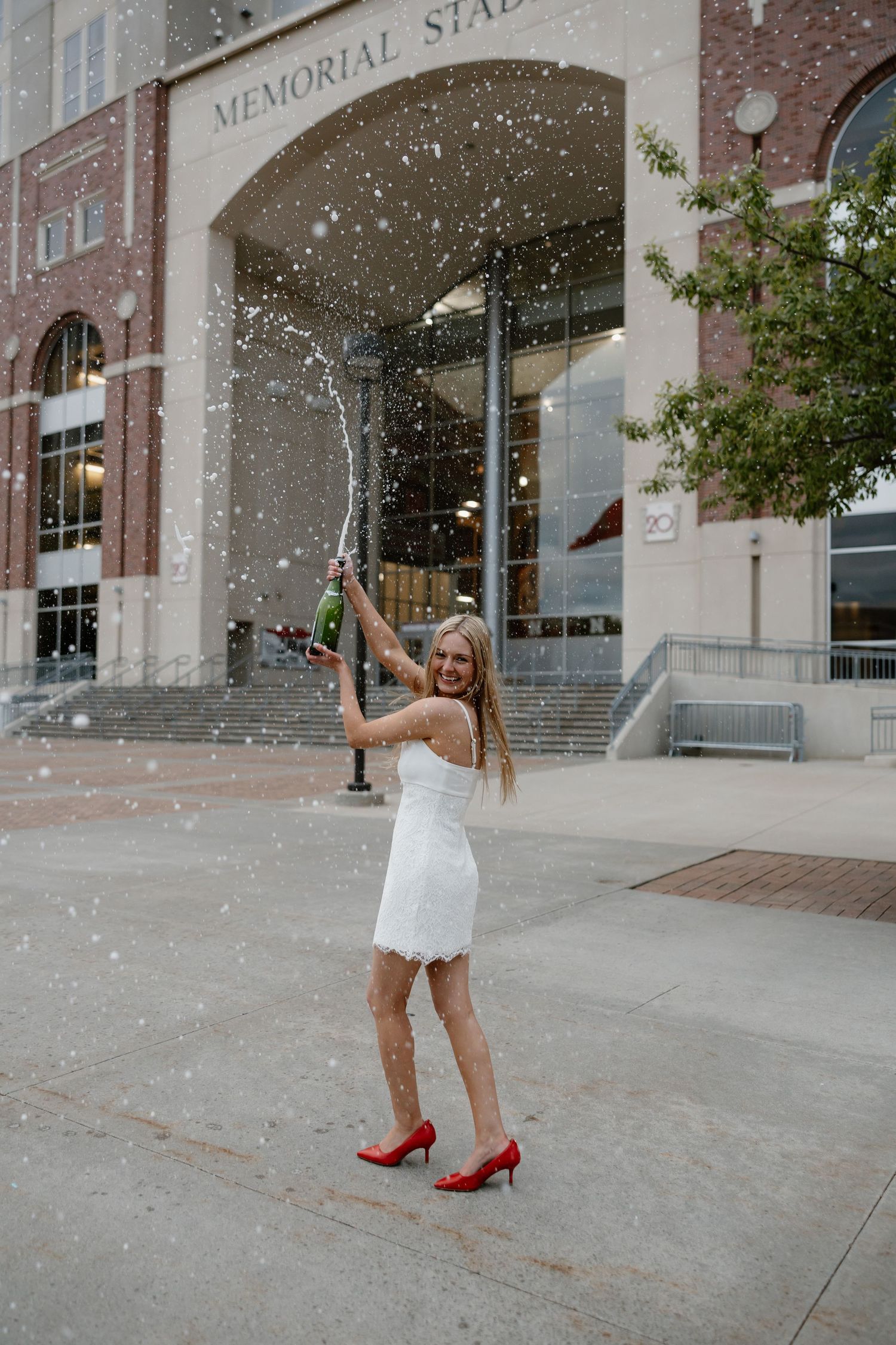 Woman in white dress and red heels celebrates with champagne outside Memorial Stadium.