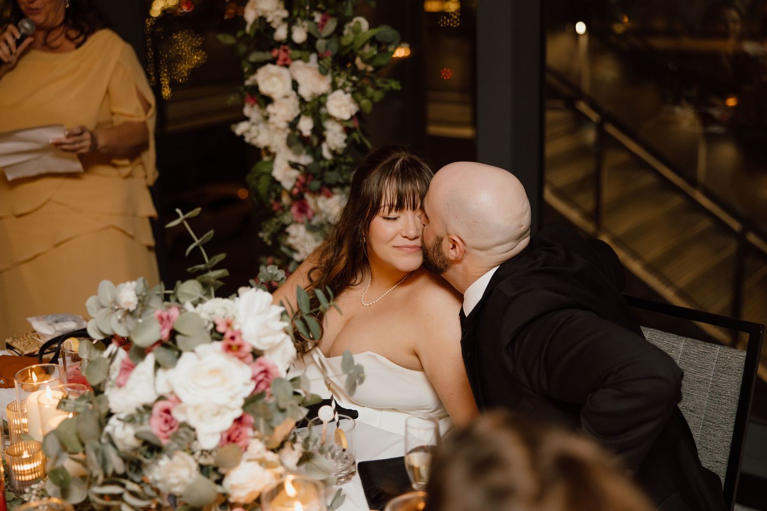 A couple shares an intimate moment at an elegant wedding reception table decorated with floral arrangements and candlelight.