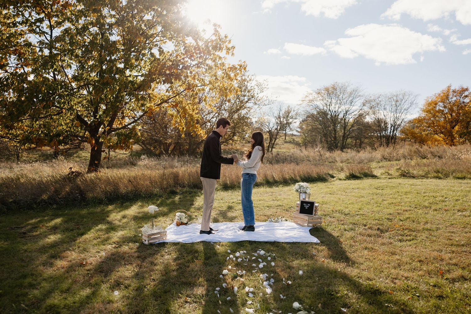 Wide angle view of a romantic proposal setup in a sunny autumn field with picnic blanket.