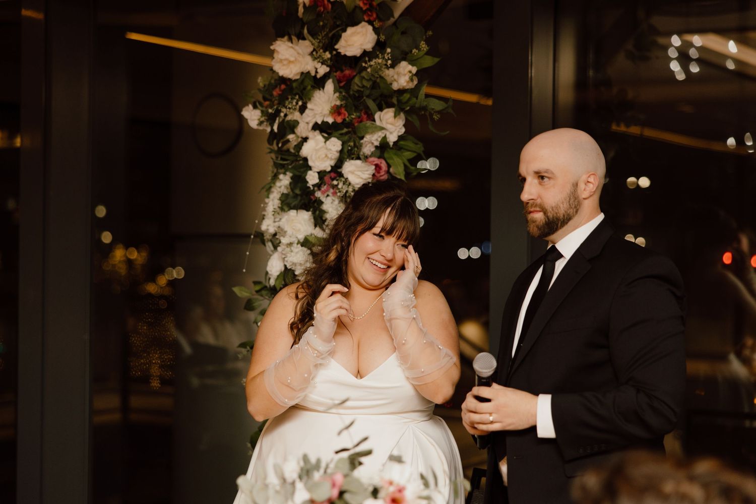 A laughing couple shares a moment during their indoor wedding reception under floral decor and dim lighting.