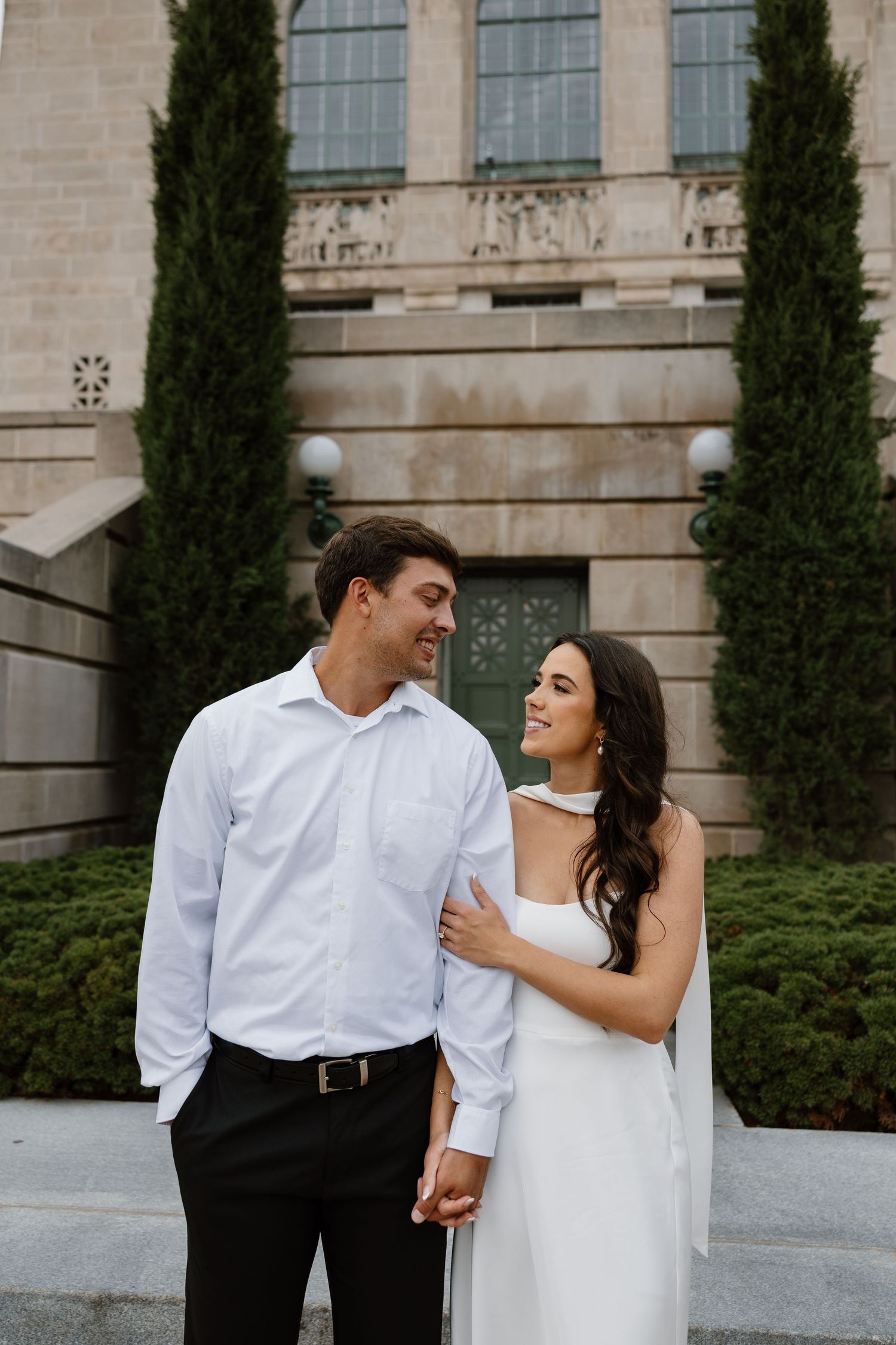 Elegant couple in white and black attire posing against stone building with cypress trees.