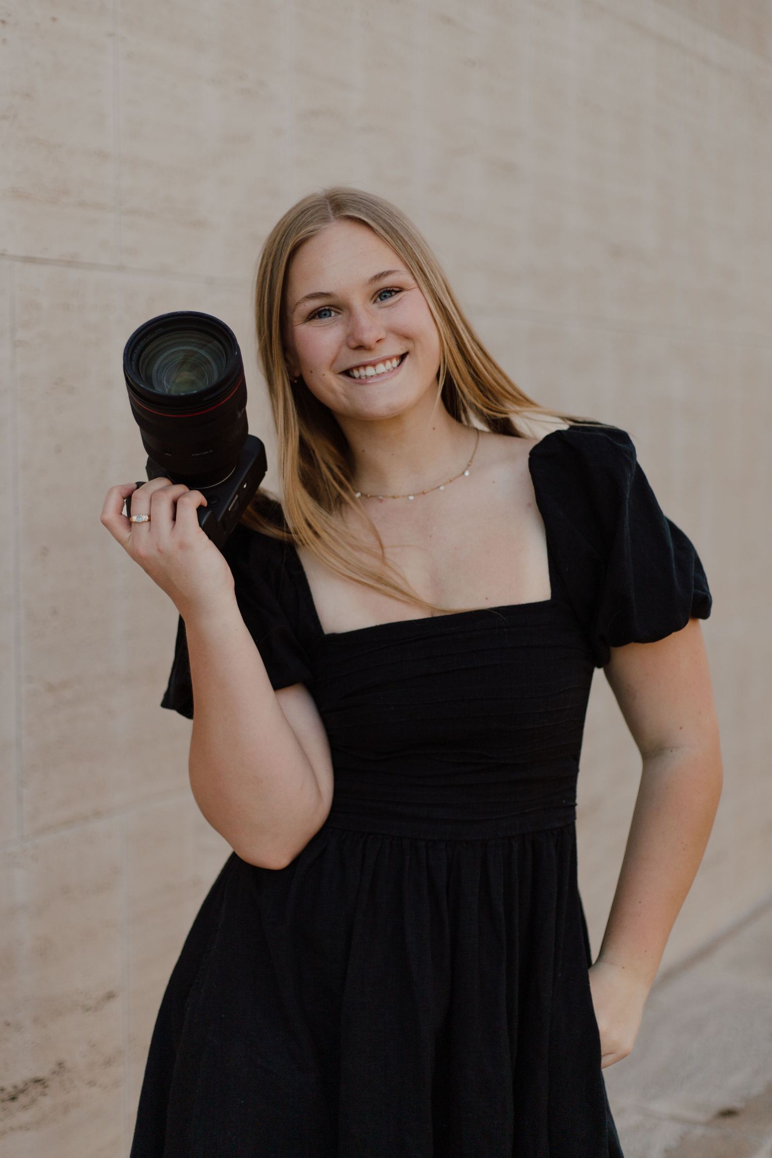Woman in black dress posing against neutral wall background.