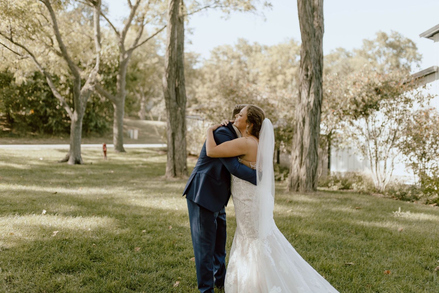 Two people embracing in an outdoor ceremony setting with tall trees and sunlit grass in the background.