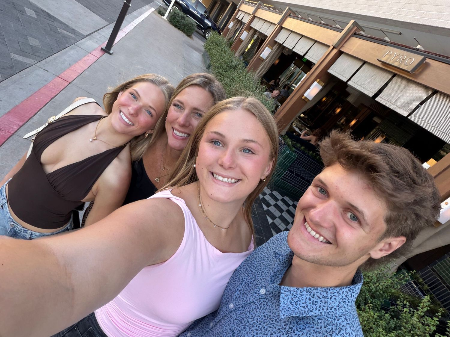 Four smiling friends taking a selfie outdoors near a restaurant patio on a sunny day.
