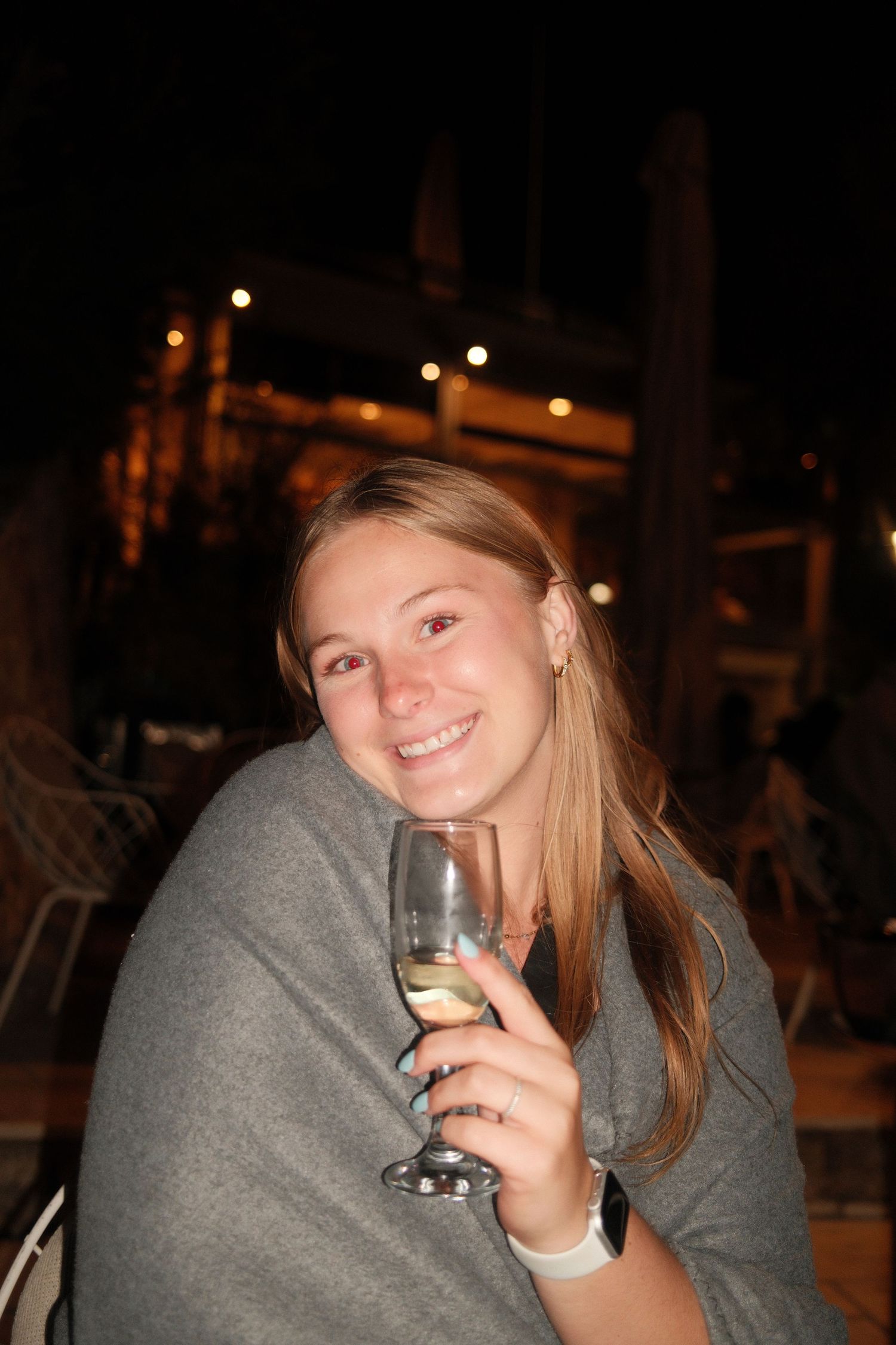 Smiling young woman wrapped in gray blanket holding white wine glass at outdoor evening venue.