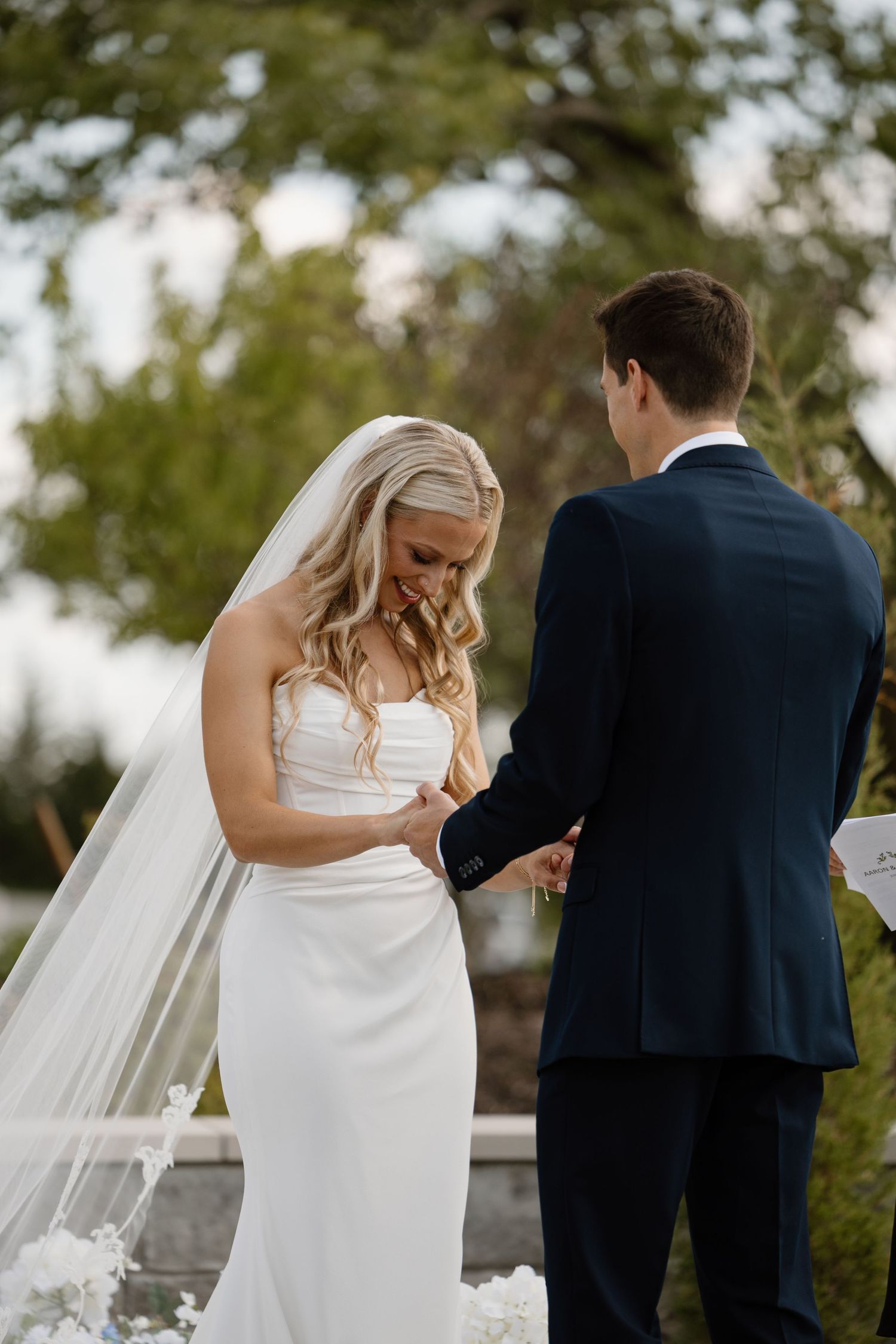 Bride in white strapless gown and veil holds grooms hands during outdoor wedding ceremony.