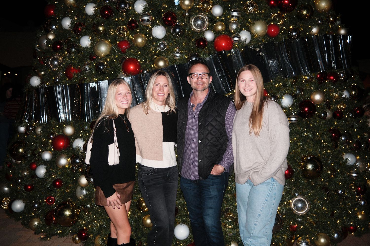 Group of friends posing in front of decorated Christmas trees with twinkling lights and ornaments.