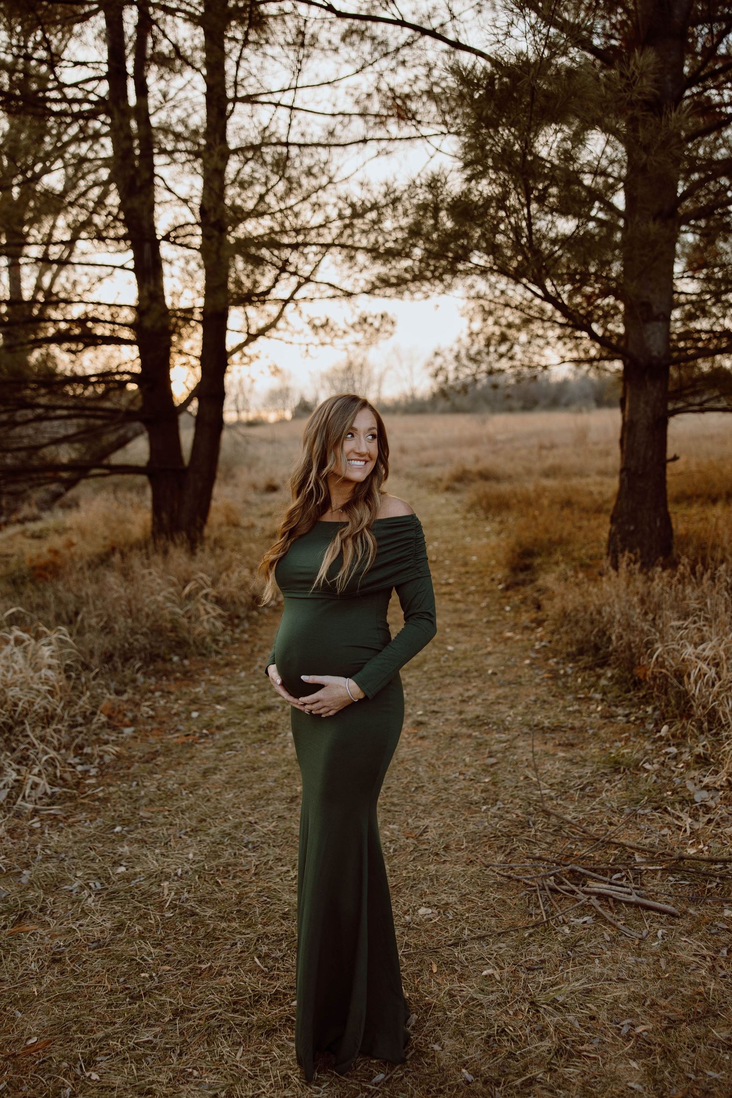 Expectant mother in forest clearing poses in flowing emerald dress at sunset.