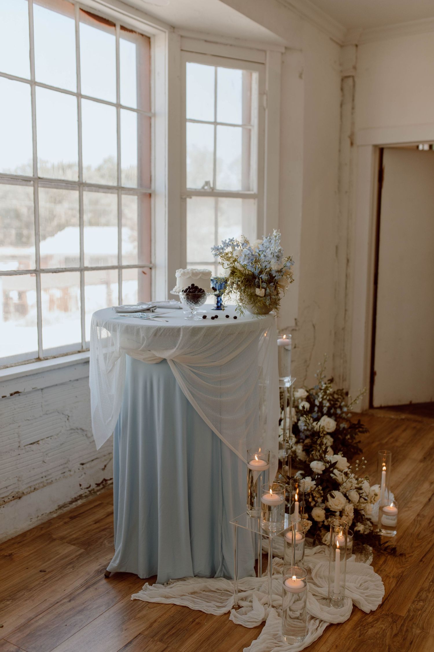 Romantic wedding decor setup with draped table, candles and floral arrangements near bright windows in rustic room.
