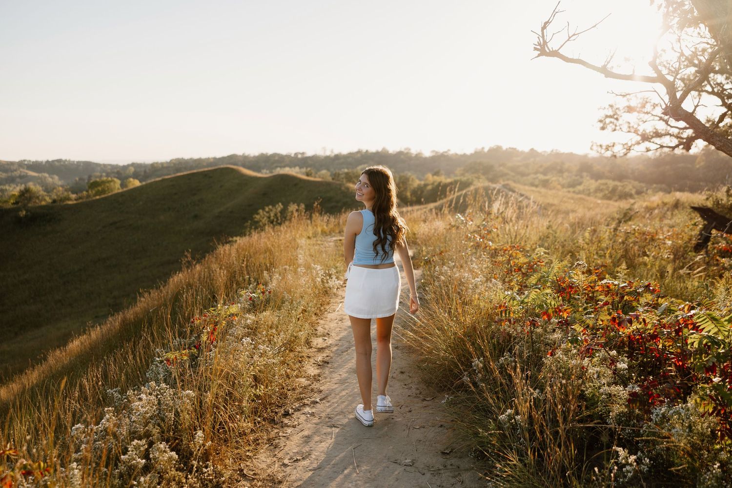 Golden sunlit trail through rolling hills with dry grass and coastal vegetation at sunset.