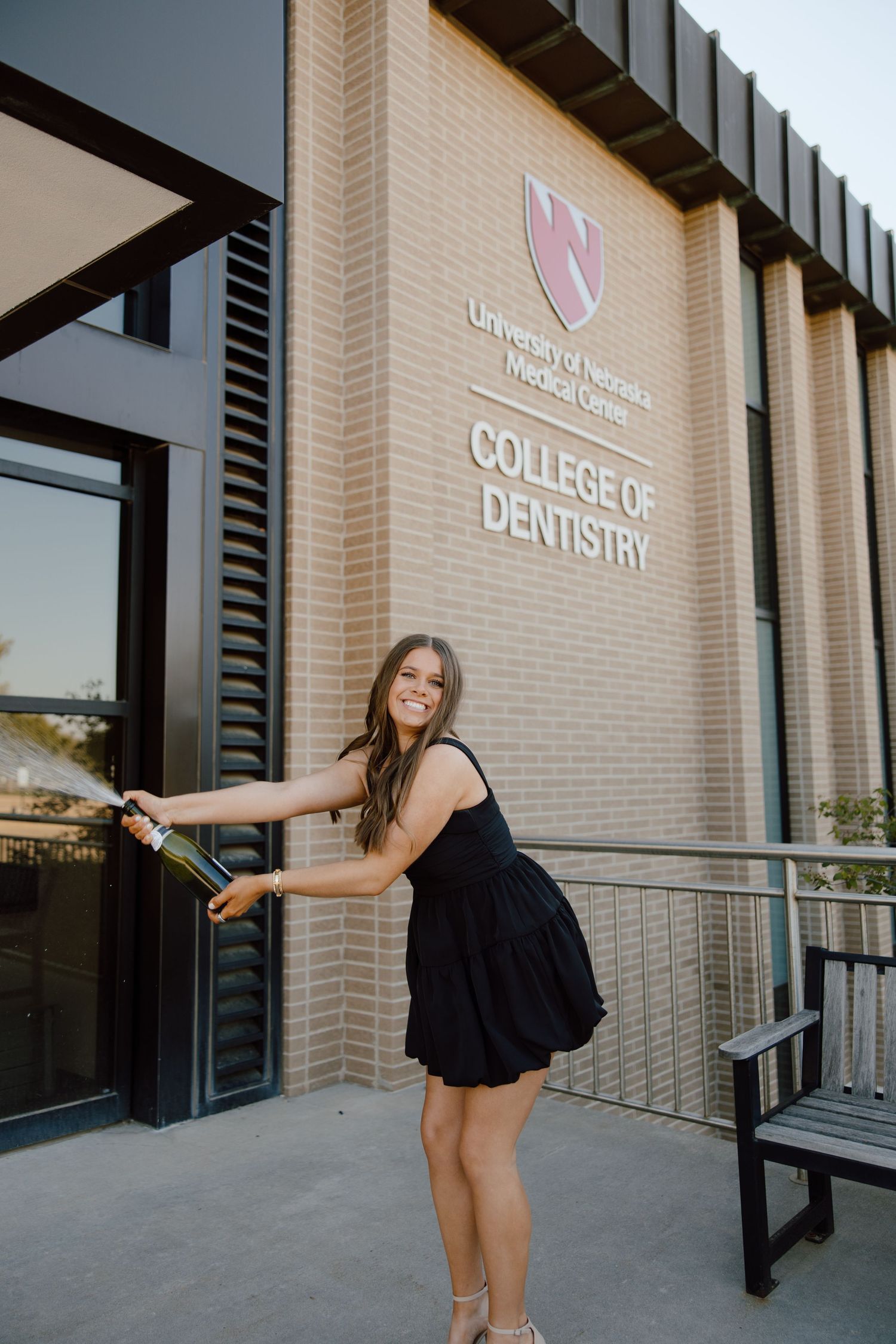 A graduate in a black dress poses playfully outside a dental school building.