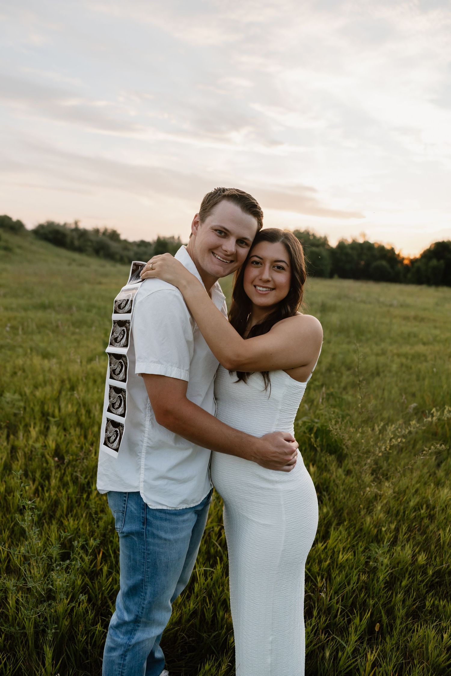 Couple embracing in sunset field while holding ultrasound photos.