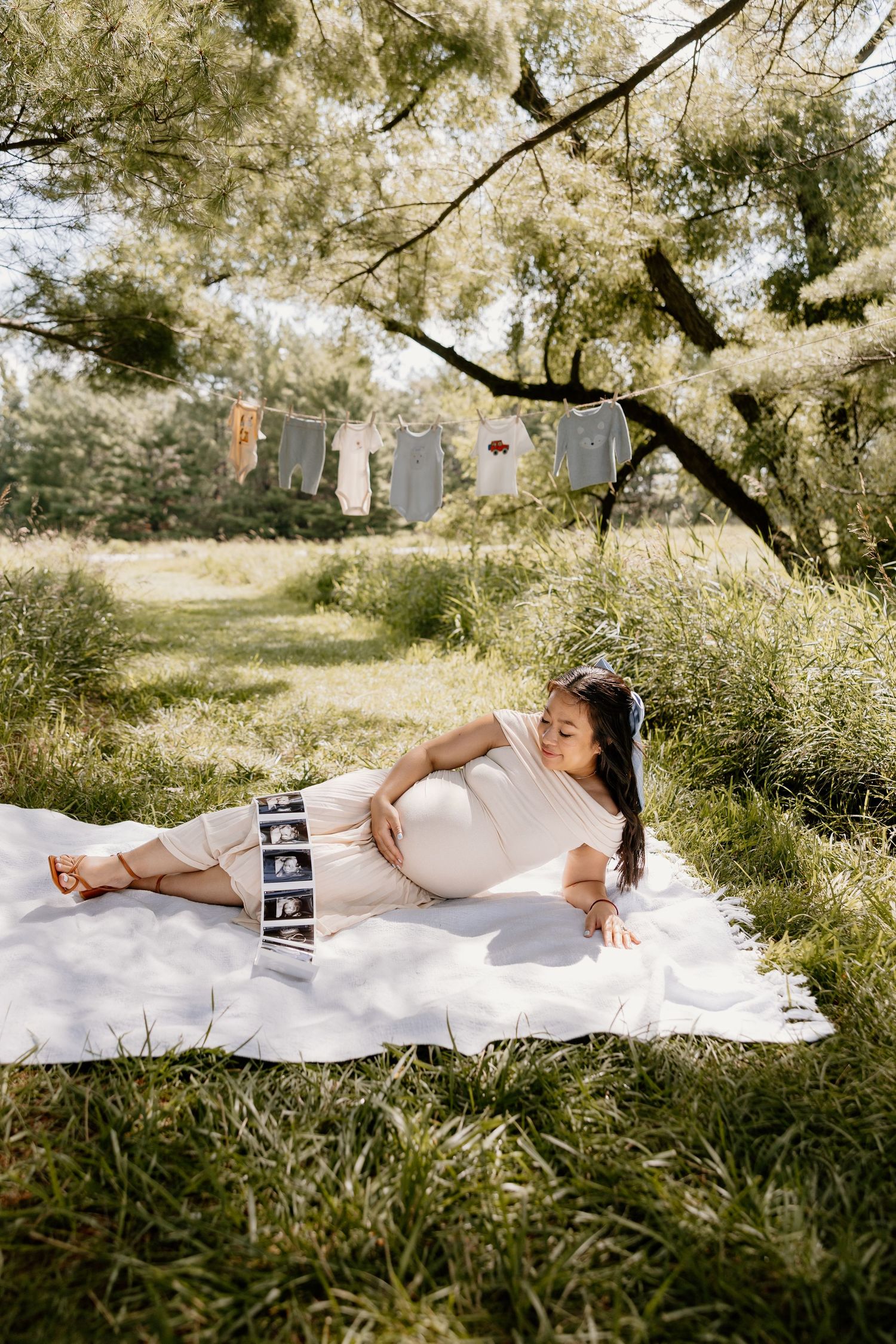 Pregnant woman in white dress lying on blanket outdoors with ultrasound photos.