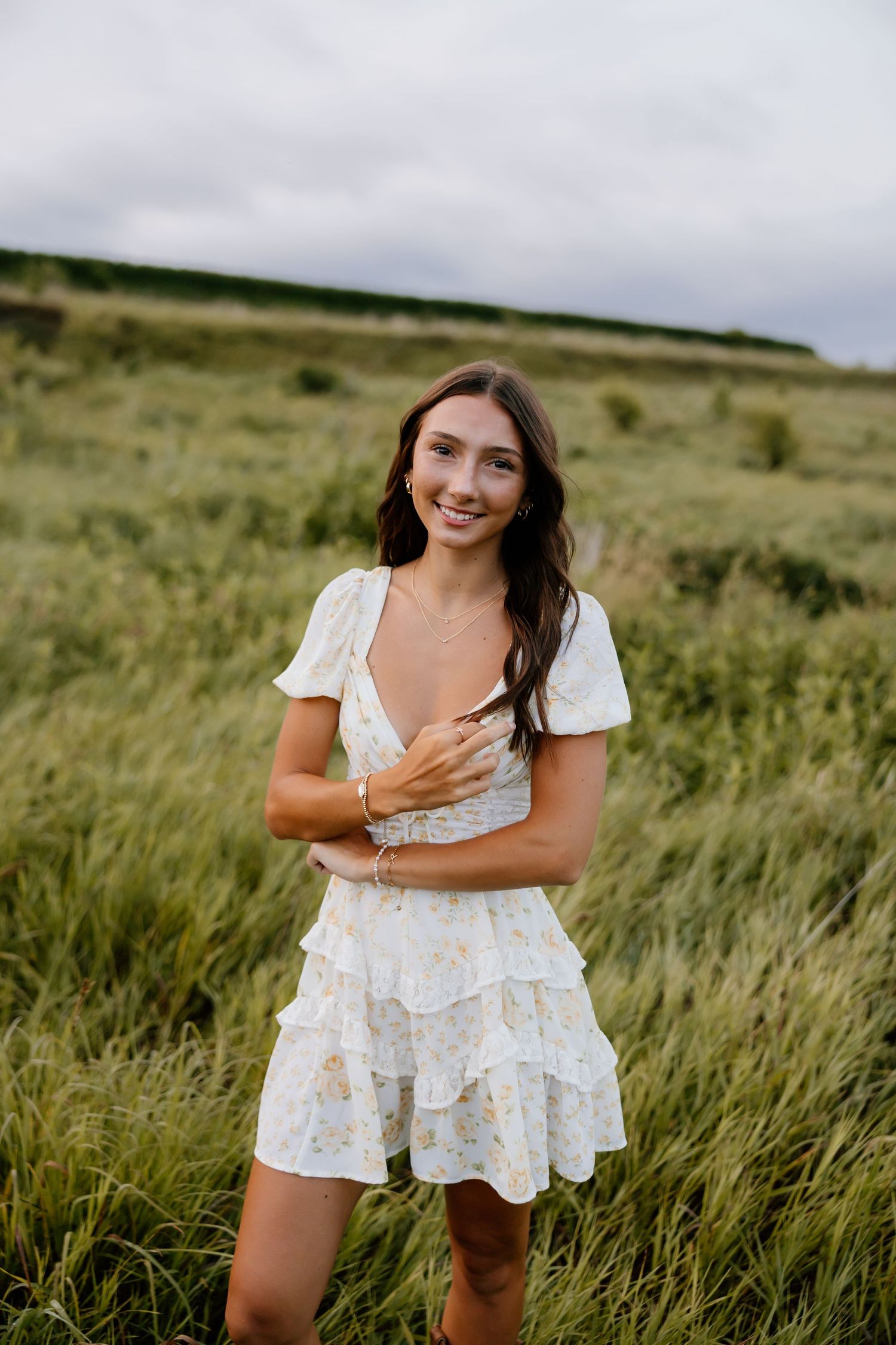 Portrait in flowing white dress amid tall grass field under cloudy sky.