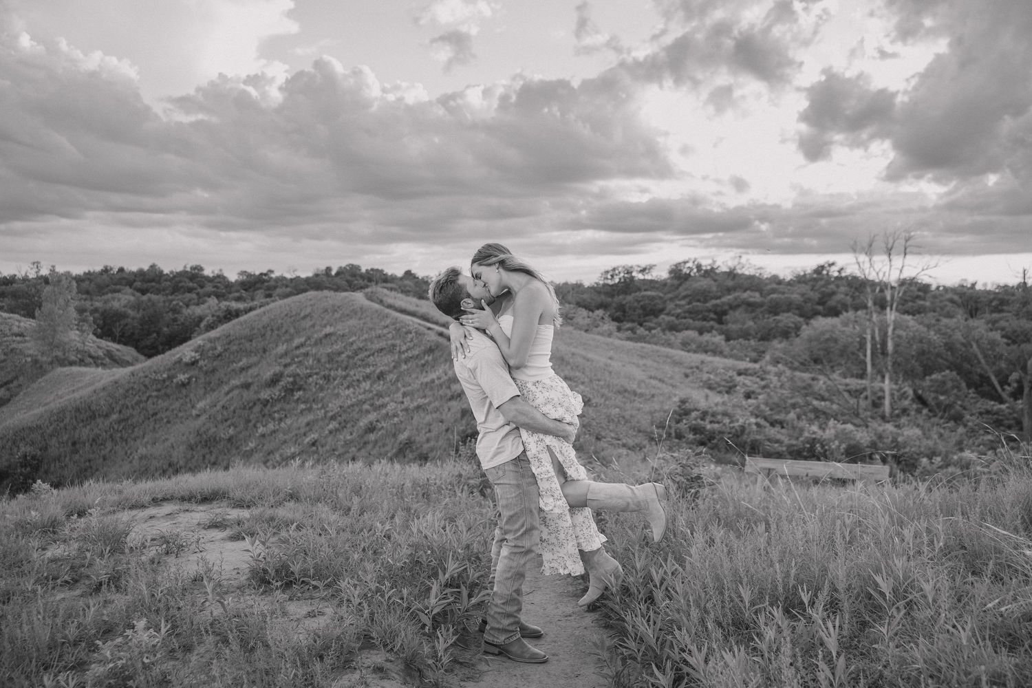 Black and white photo of a couple embracing outdoors, man lifting woman against a scenic landscape with rolling hills.