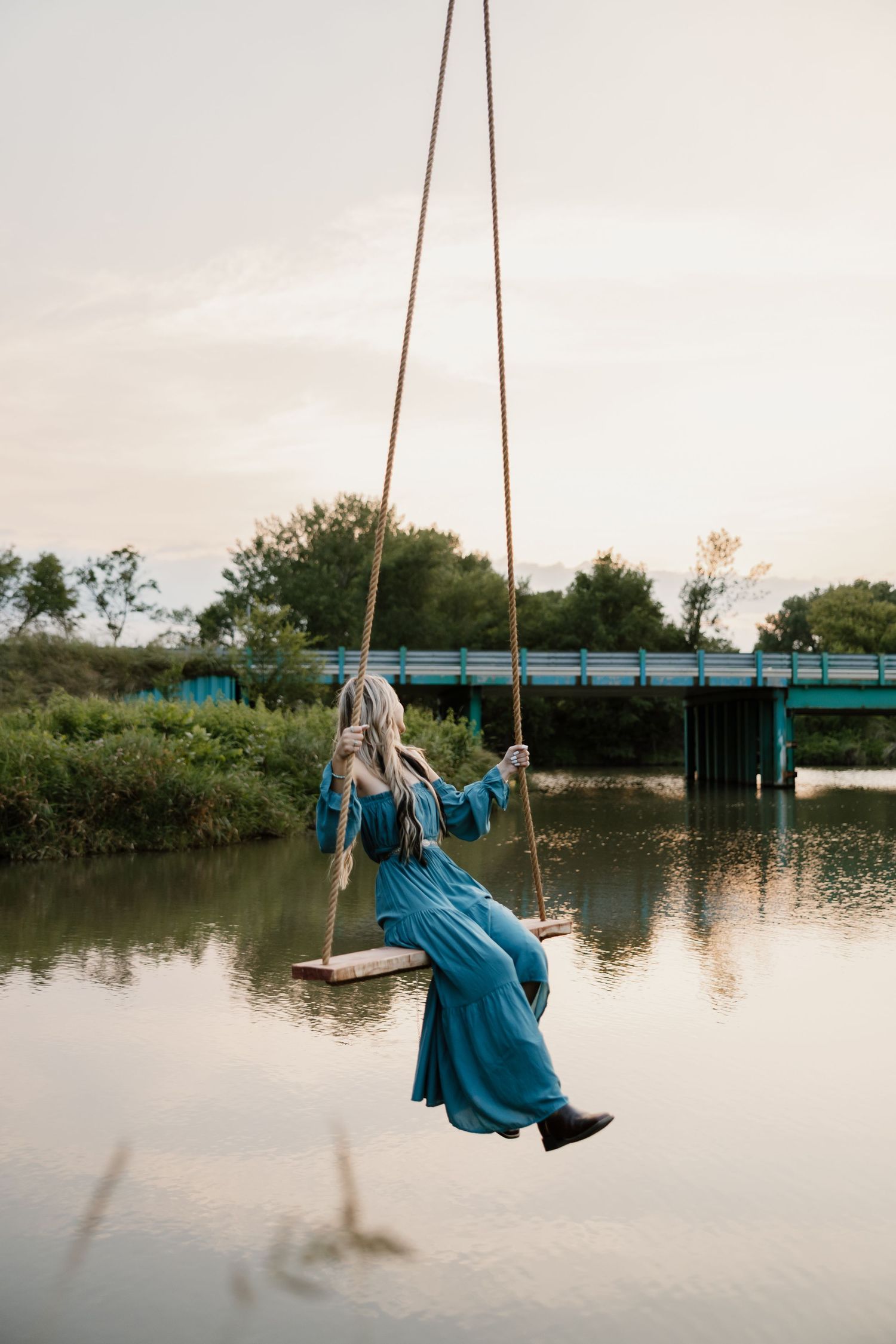 A person in a blue dress sits on a rope swing over a tranquil lake at sunset creating a peaceful scene.